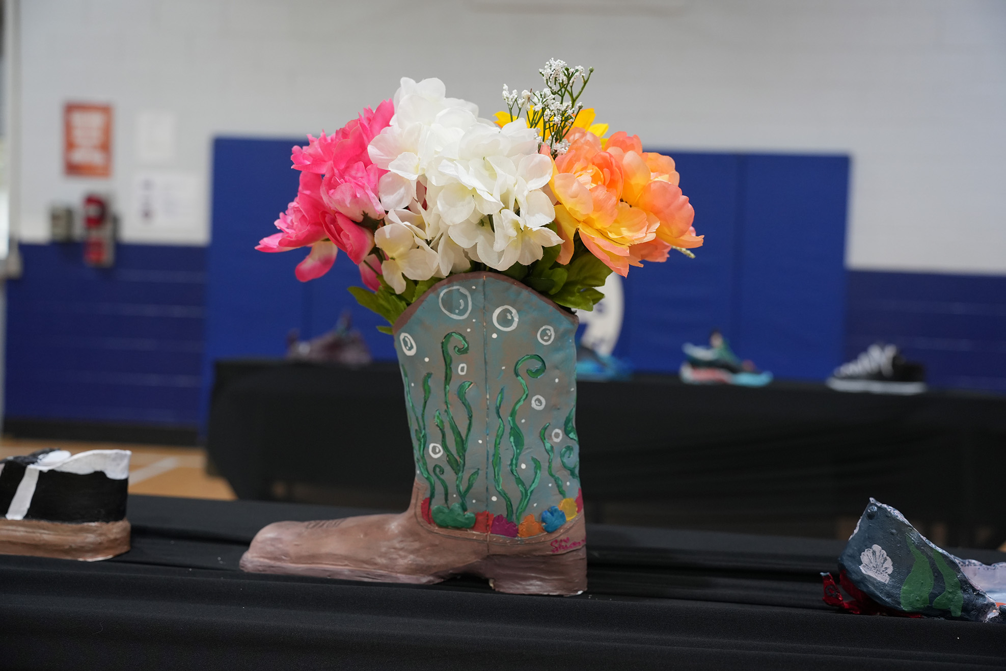 A large boot decorated with flowers sits on a black table in a gym with blue walls.