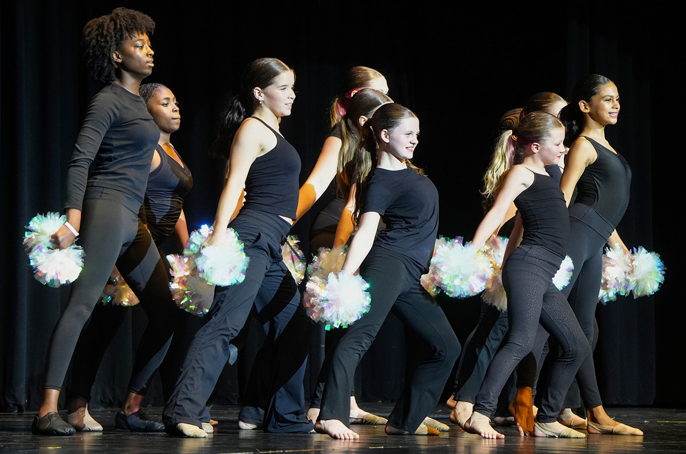A group of young women perform a routine on stage with pom-poms, dressed in black attire.