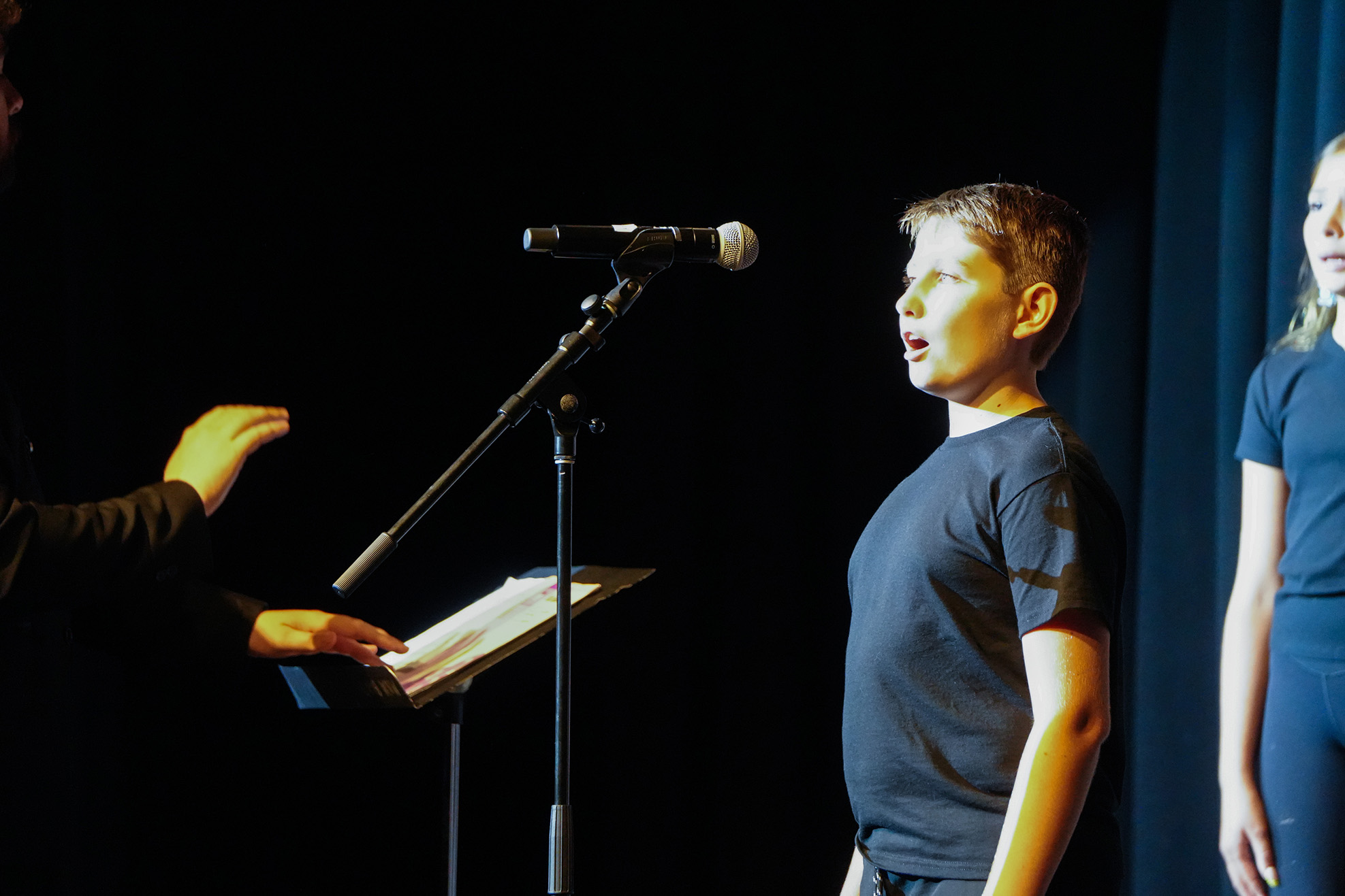 A boy sings on stage, holding a microphone, while another person holds a music stand nearby. A girl stands behind him.