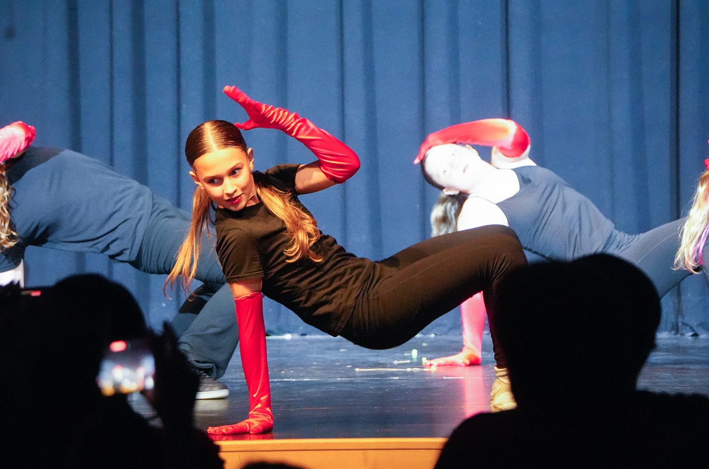 Dancers in red gloves perform on a stage with blue curtains. One dancer performs a split. Audience members watch from the front.