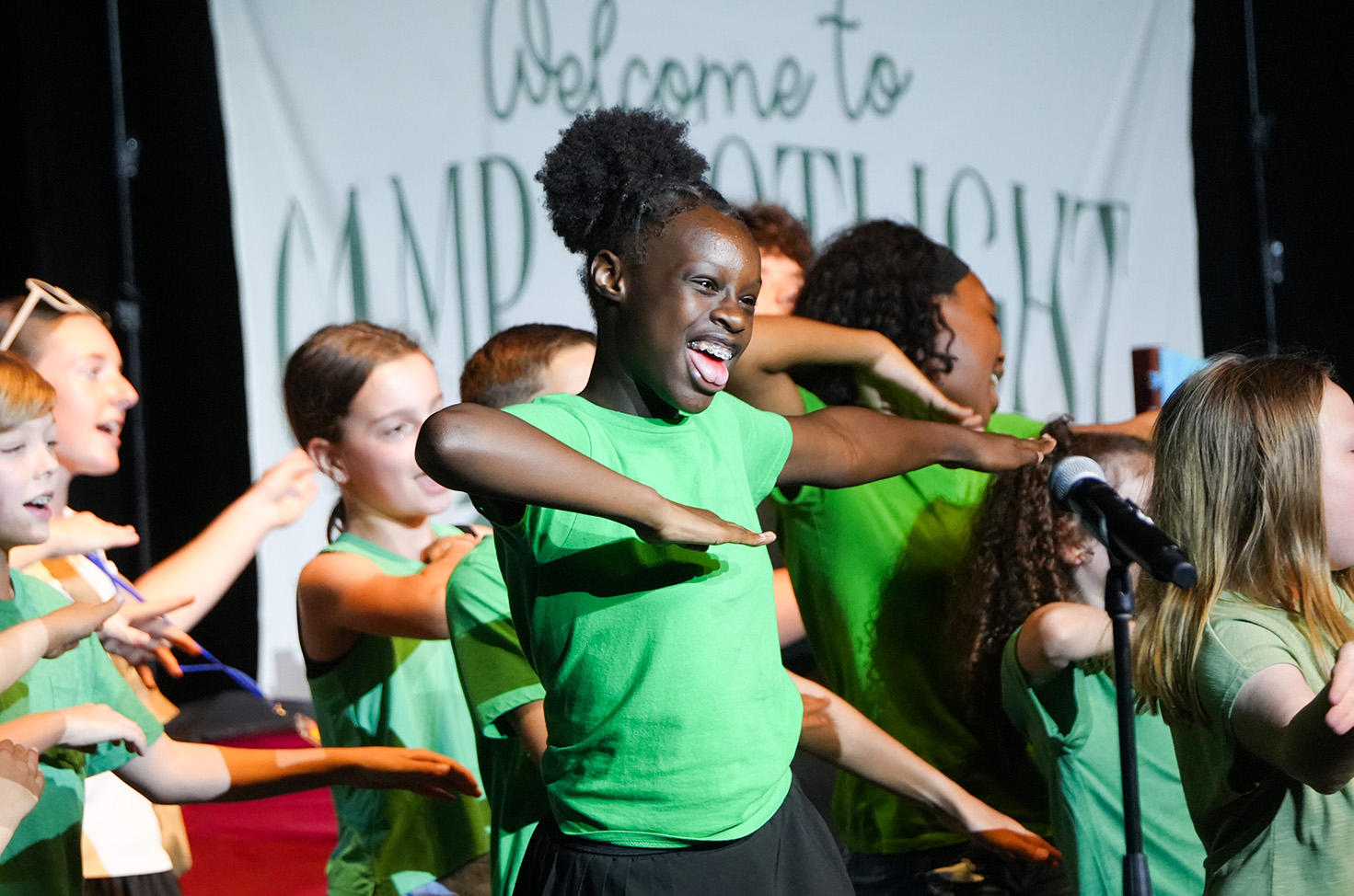 A group of children in green shirts perform on a stage, with a banner in the background.