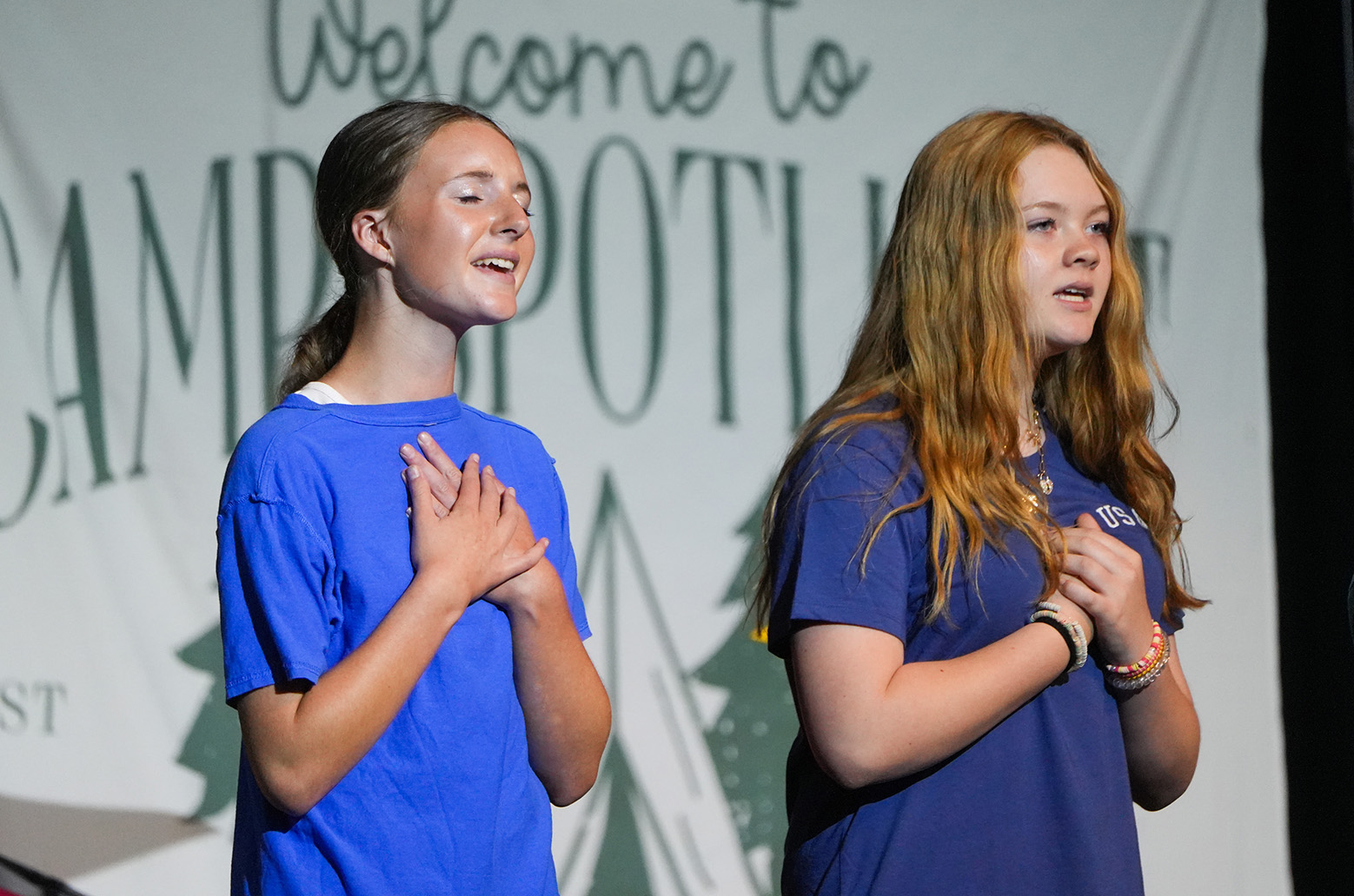 Two girls in blue shirts clasp their hands in front, standing before a banner with "Welcome to Camp Potwin" in large letters.