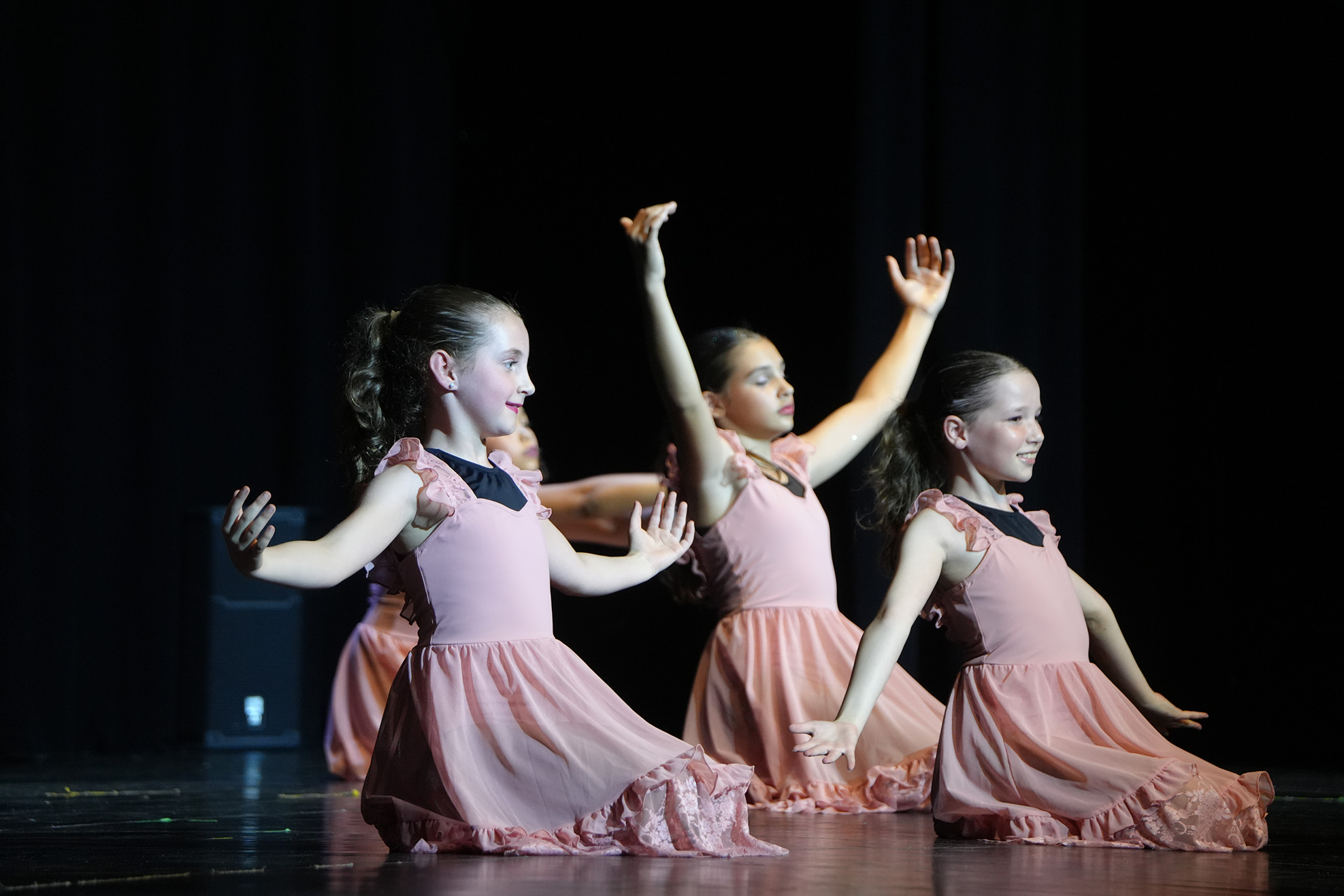 Four young girls in pink dresses perform a dance routine on a stage with dark background.