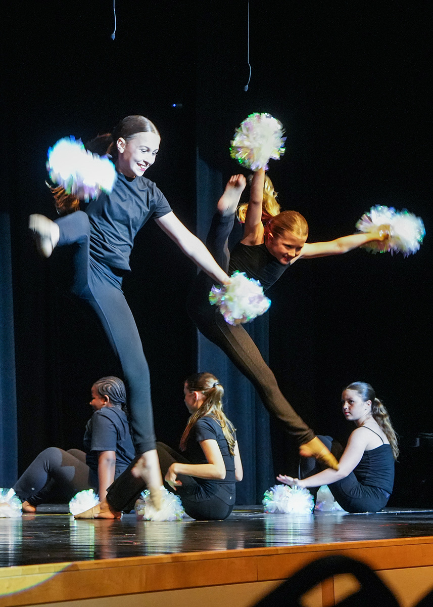 Two women in black performing on stage with pom-poms while others sit behind them.