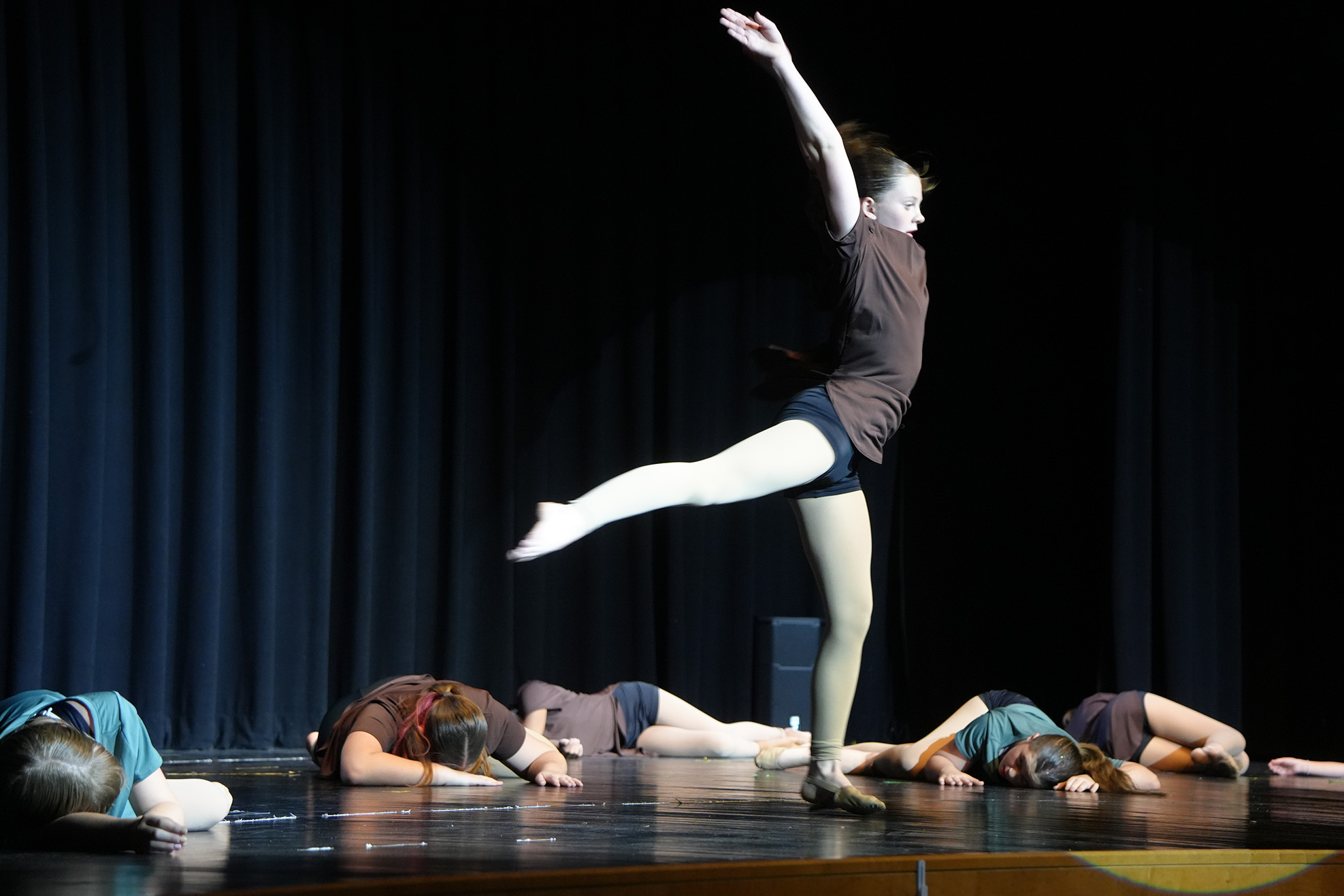 Dancer in spotlight on stage, others lying on stage, blue curtains in background.