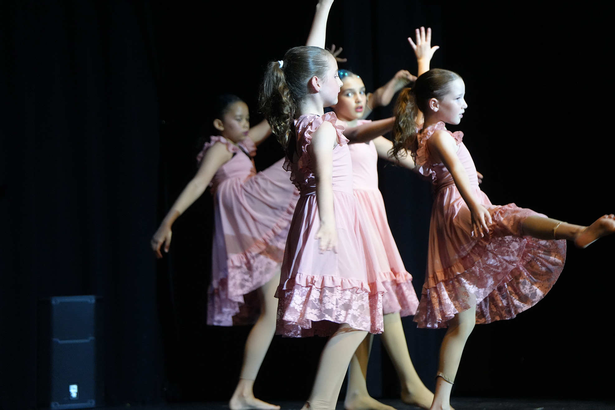 Four girls in pink dresses perform a dance on a stage with a black curtain backdrop.