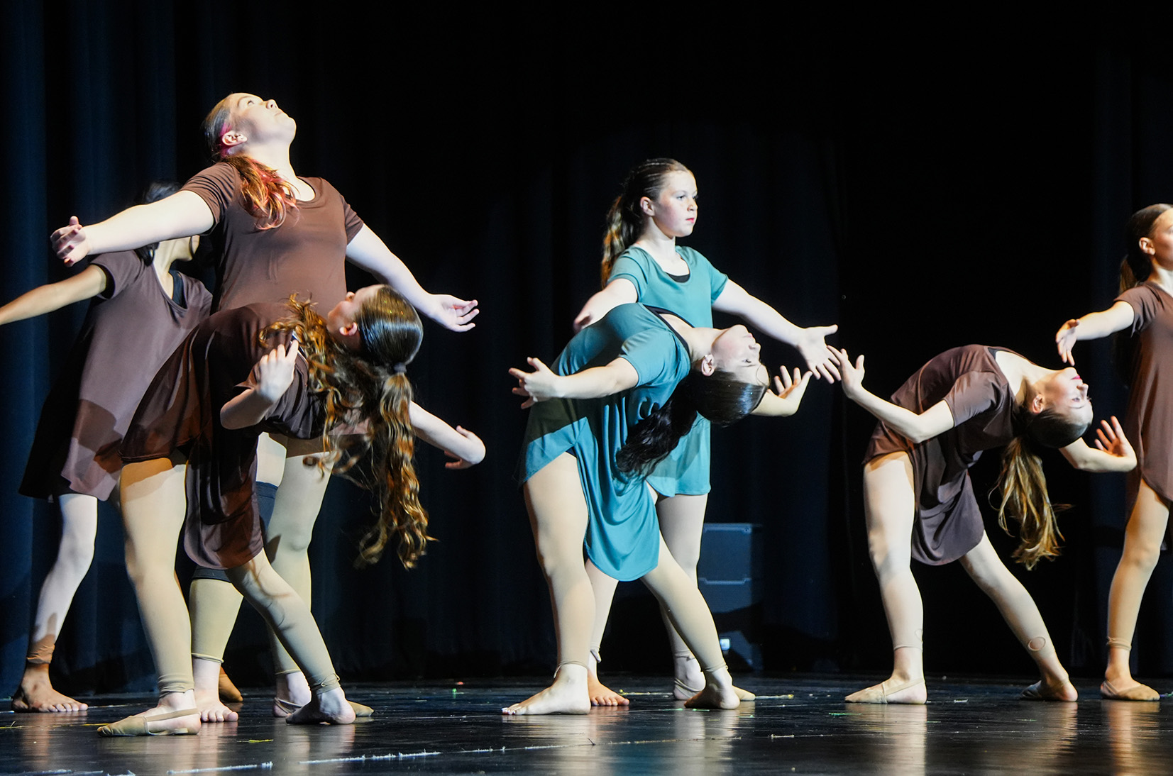 Women in various outfits perform a dance routine on a stage with black curtains in the background.