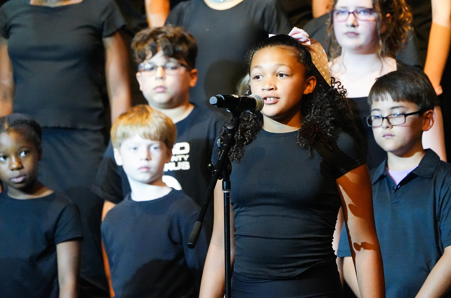 Group of children, mostly wearing black shirts, standing on stage. One child sings into a microphone.