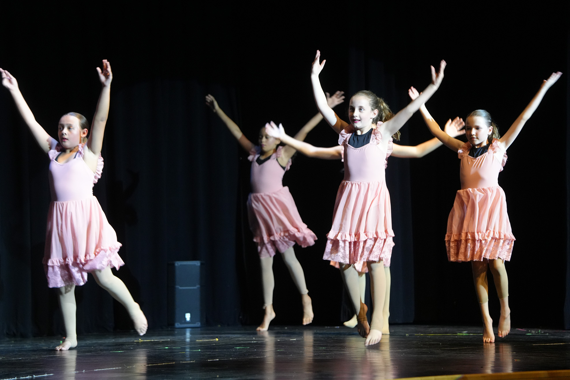 Five young girls in pink dresses perform a dance routine on a stage with a black background.