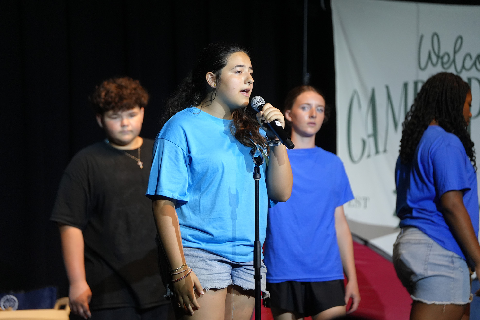 Four people in light blue shirts, one speaking into a microphone, on a stage with a banner.