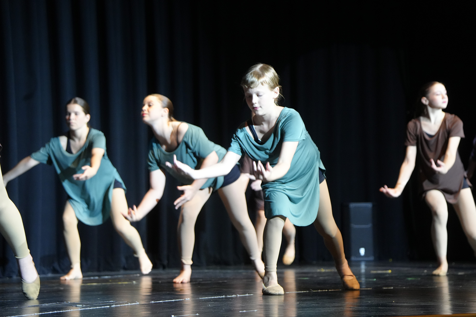 Five women dance on stage wearing various colored dresses and ballet shoes. One woman wears a brown top.