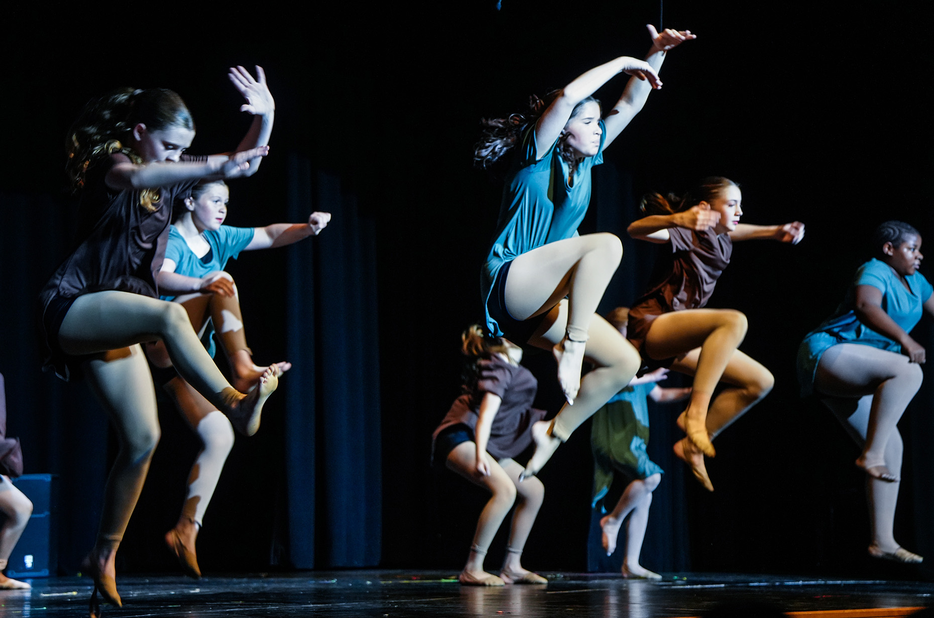 Performers in matching blue shirts and tights execute high jumps and kicks on a stage with a dark background.