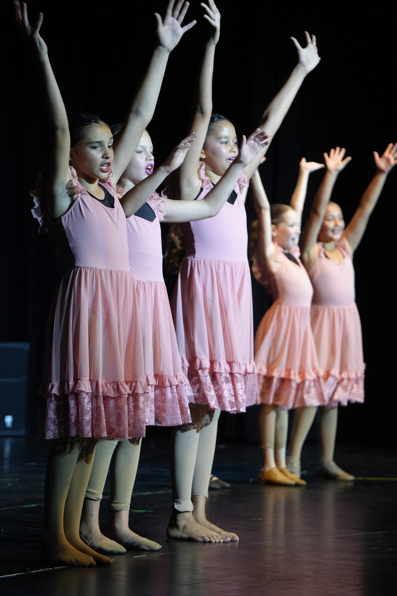 Five girls in pink dresses perform on stage, raising their arms. They wear ballet shoes and are illuminated by stage lights.