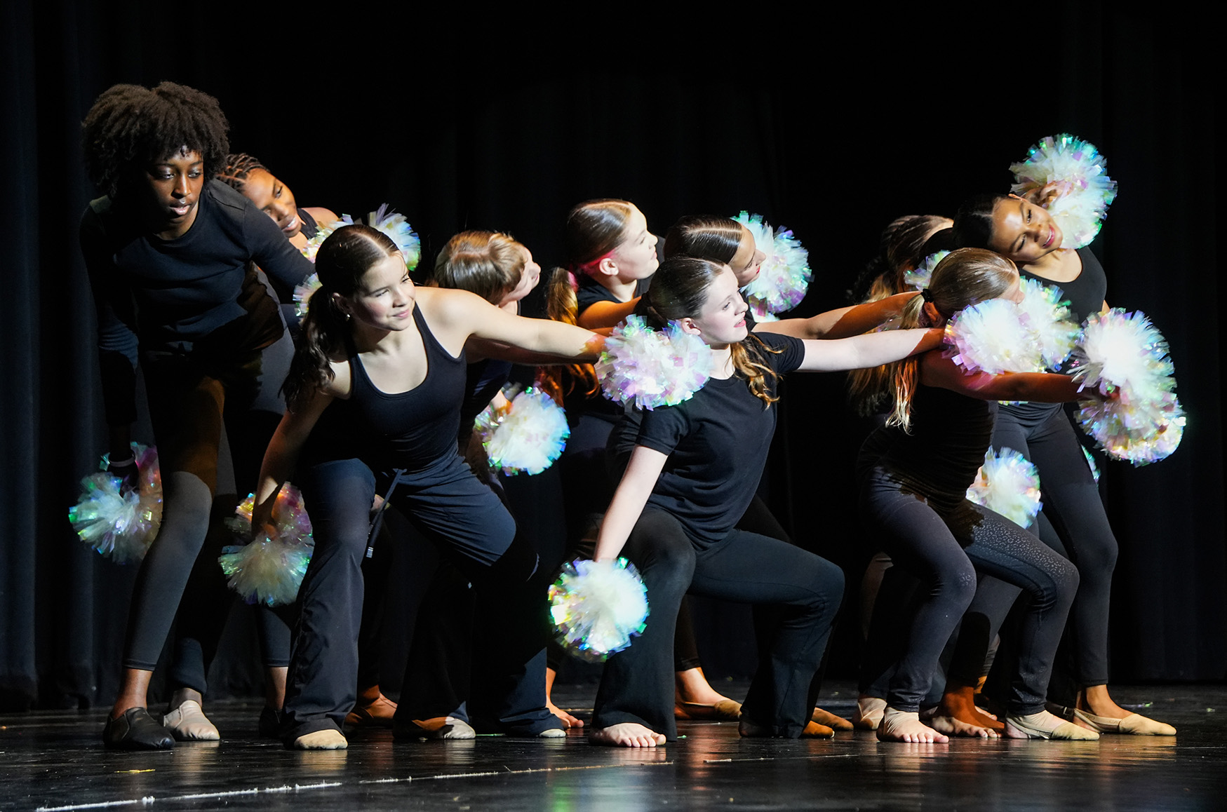 Performers on a stage in black outfits and sneakers hold pom-poms in their hands.