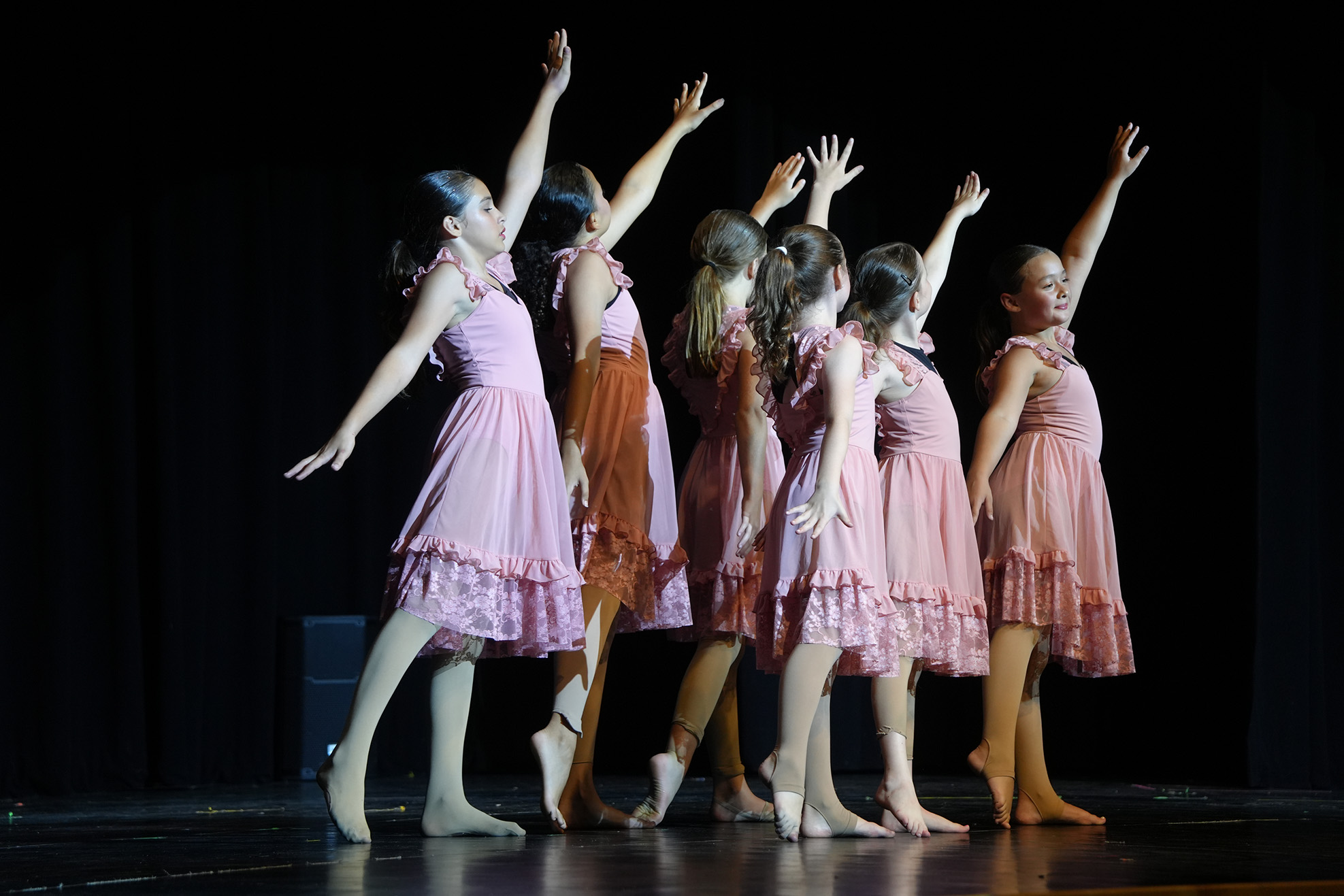 Girls in pink dresses perform a dance routine on a stage, with their arms raised.