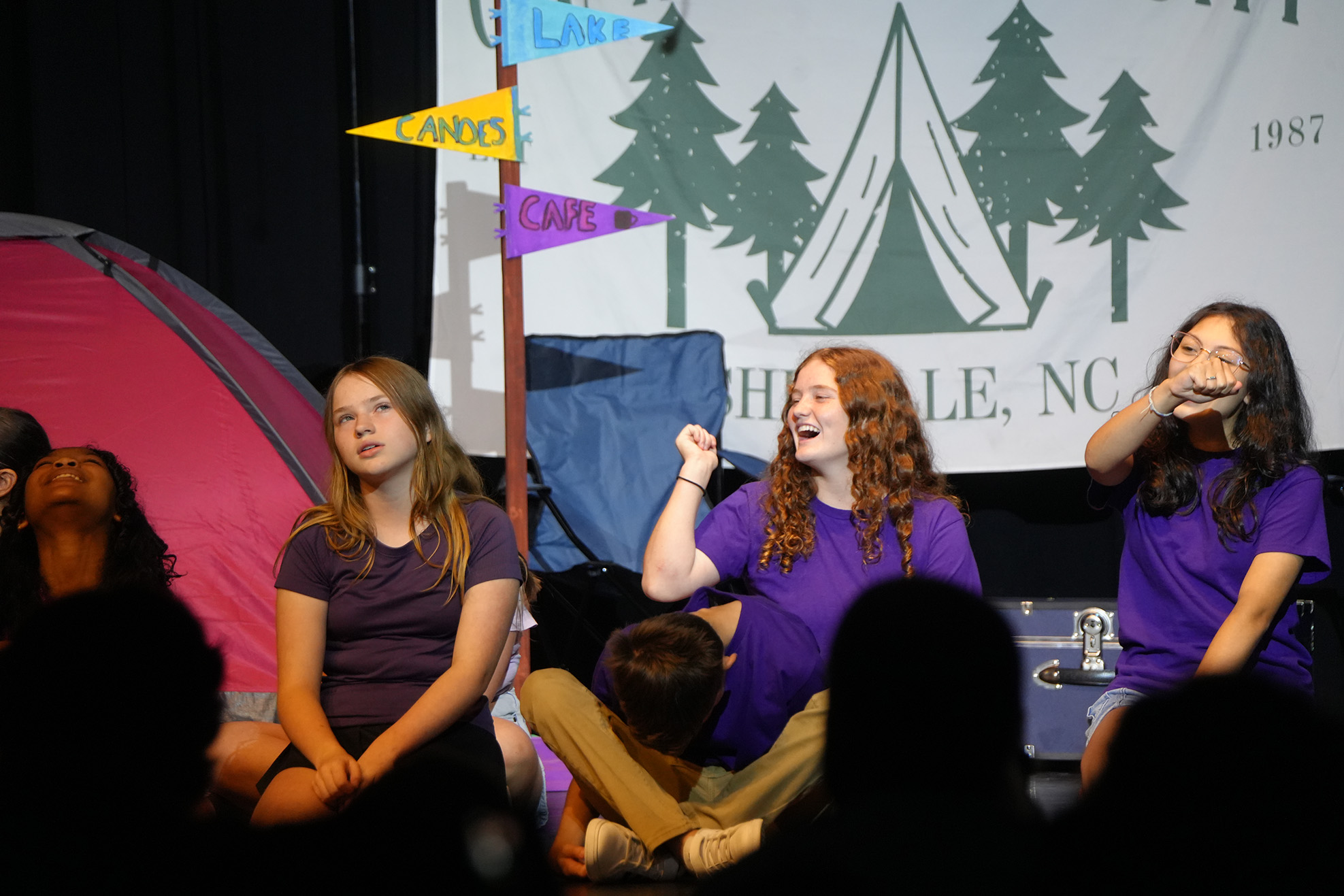 Four children in purple shirts sit on stage with a tent and banners in the background.