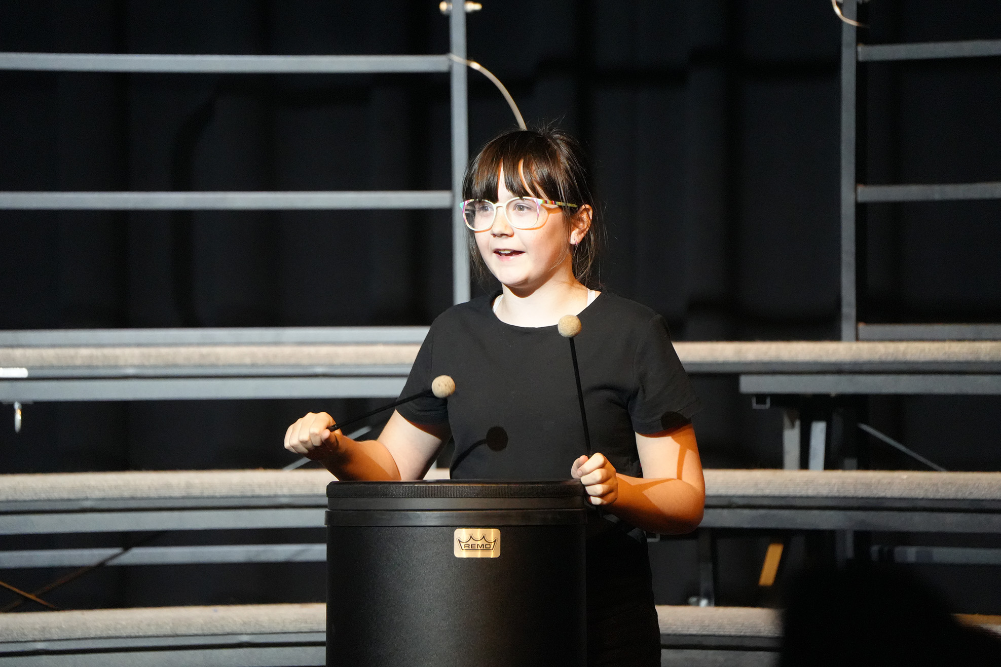 A young person in a black shirt and glasses plays drums on stage, with empty bleachers in the background.