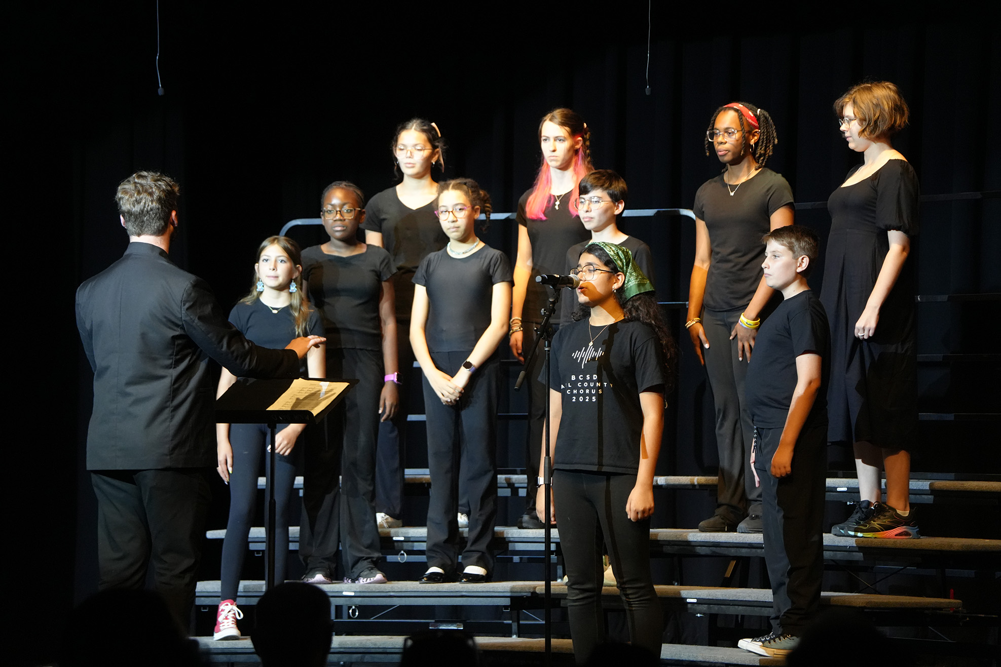 A choir of children stands on stage, with a conductor in the foreground. The children are wearing black clothing.