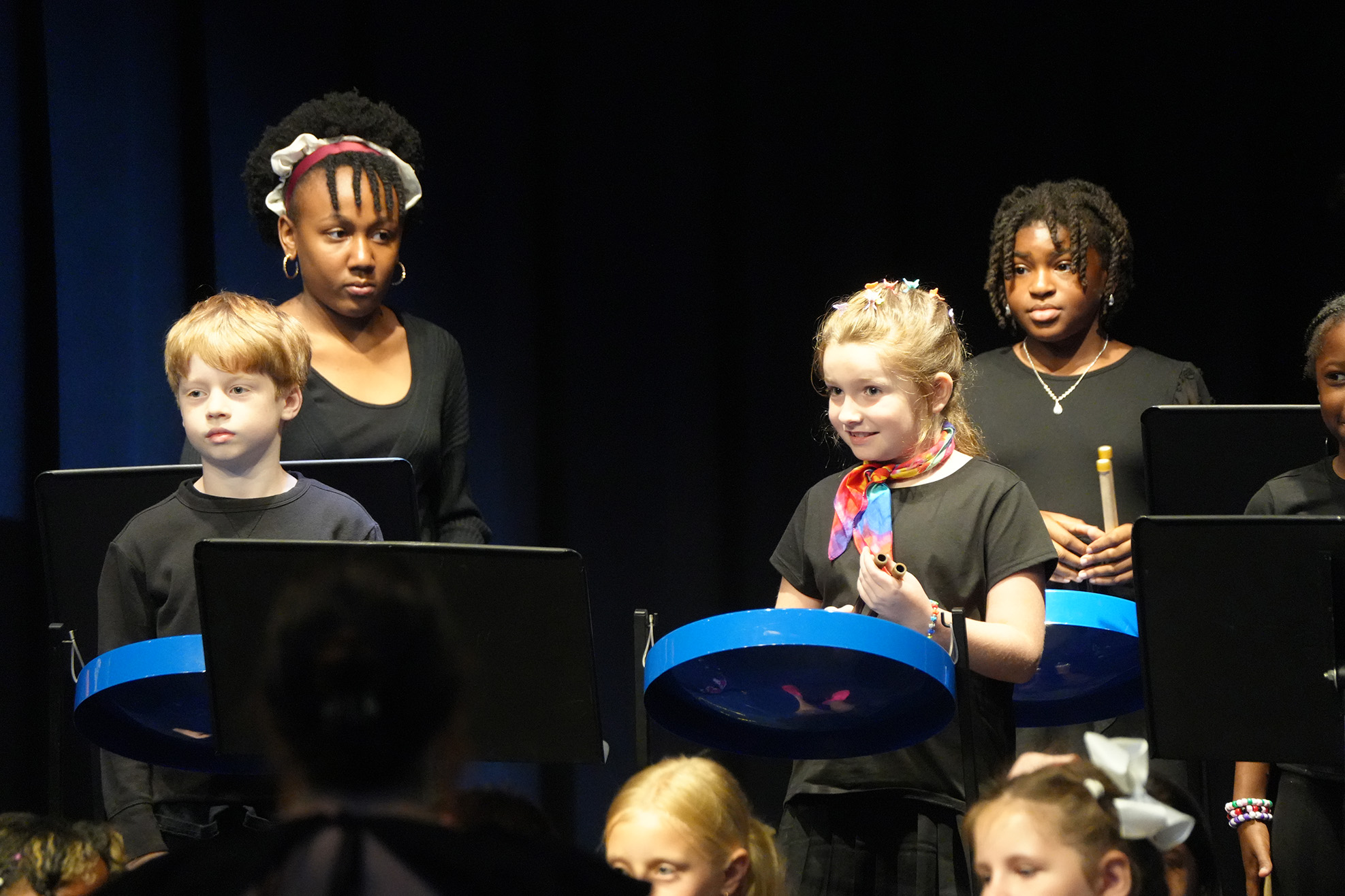 A group of children in black shirts on a stage, playing instruments. Some adults stand behind them.