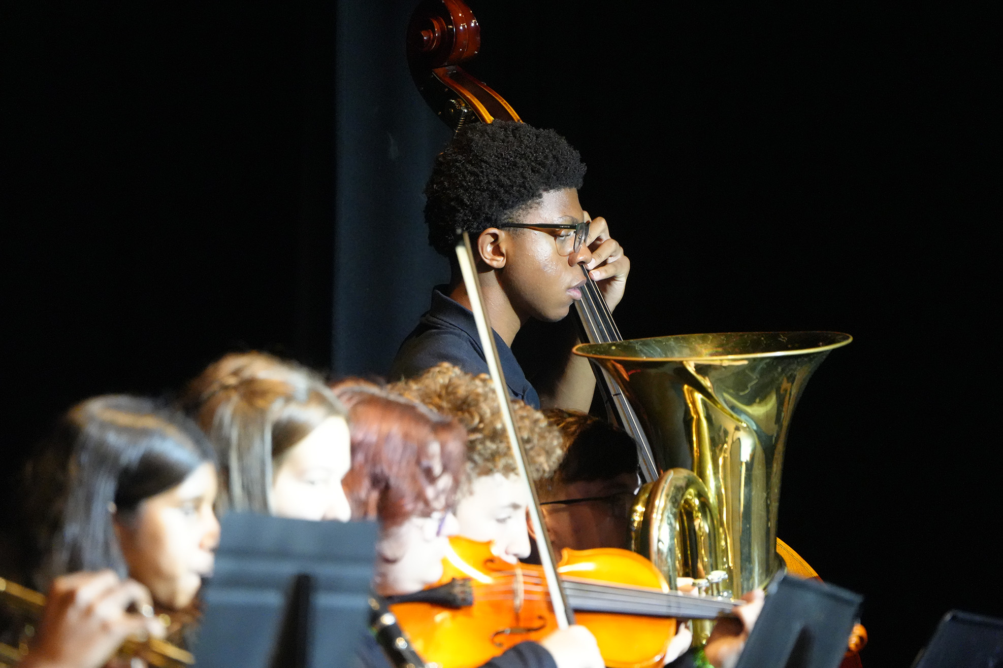 People perform in a band, including a boy on cello, a girl playing a violin, and others on trumpets and flutes.