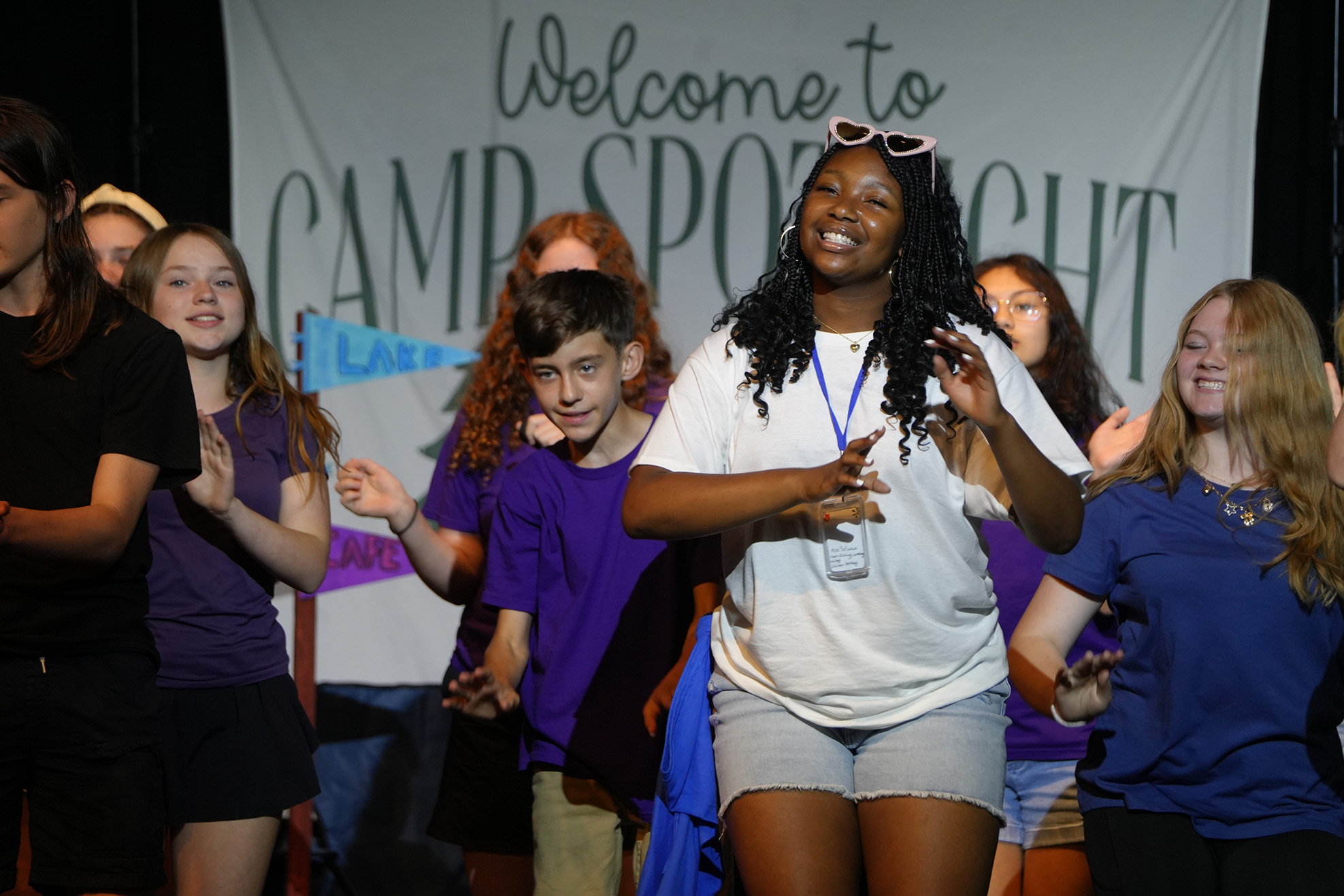 A group of young people on a stage, one woman wearing white and sunglasses gestures. Behind them is a banner that reads "Welcome to CAMP SPOTLIGHT".