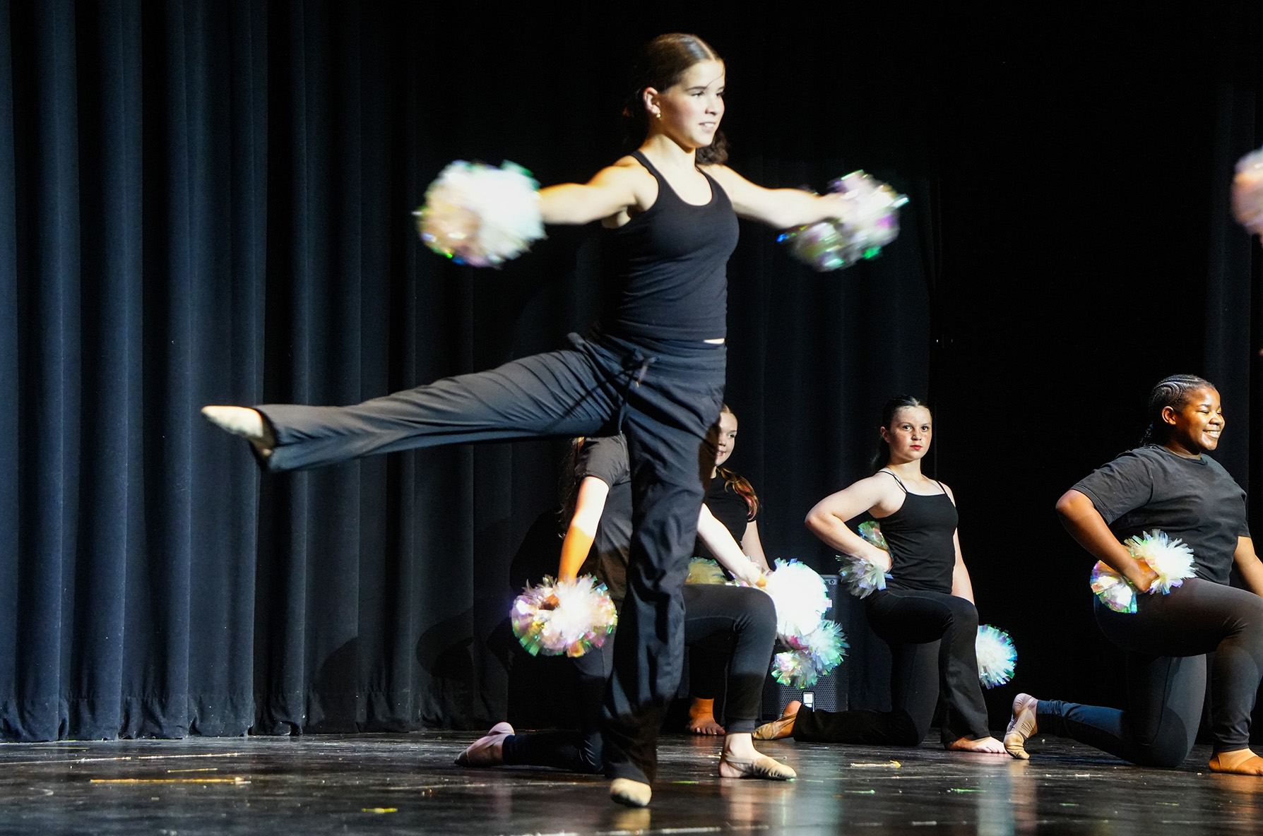 Dancers performing on a stage, one in mid-air with pom-poms, others kneeling behind her.