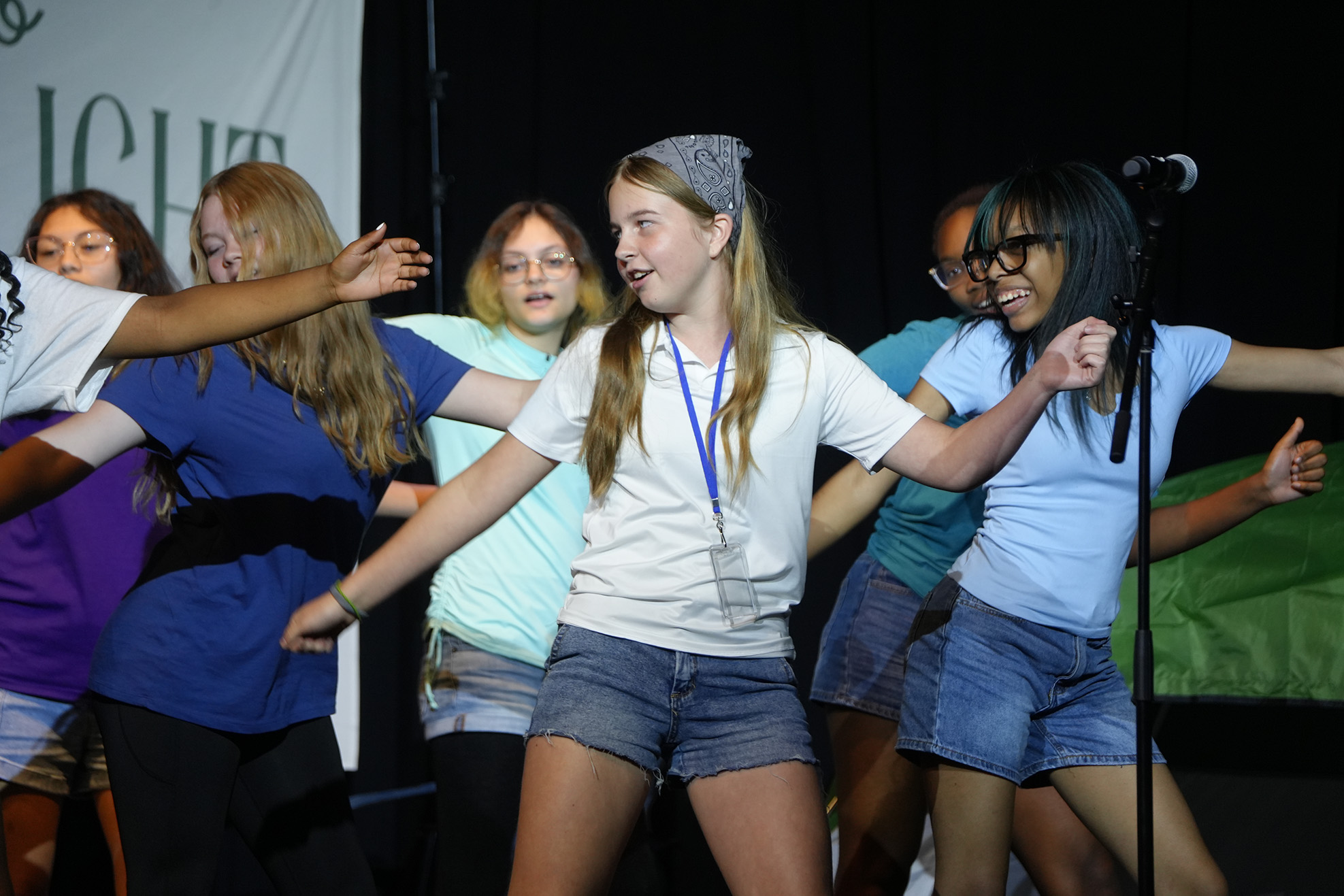 On stage, a group of young people dance with arms outstretched. One wears a white shirt, denim shorts, and a bandana.