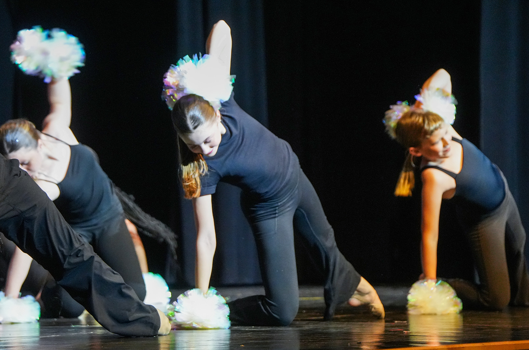 Four performers on stage with colorful pom-poms, two kneeling and two standing. They wear matching dark outfits.