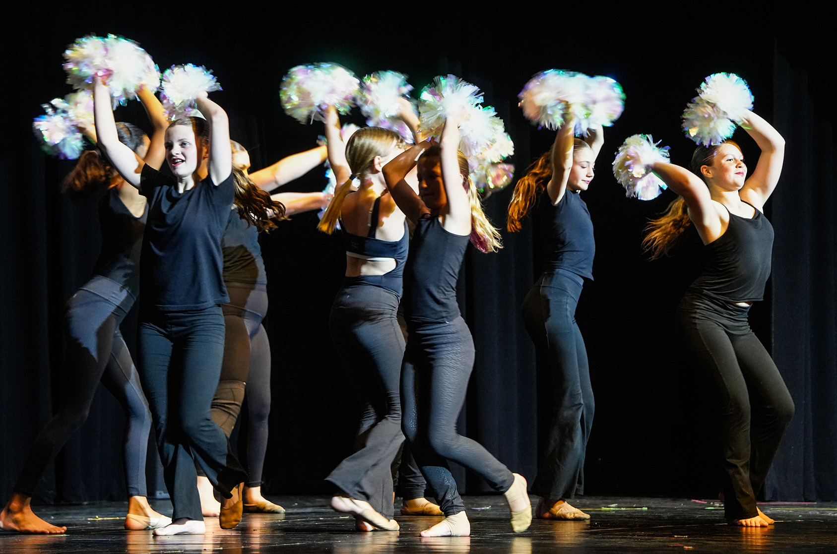 A group of women on stage wearing black tank tops and leggings, holding white and purple pom-poms.
