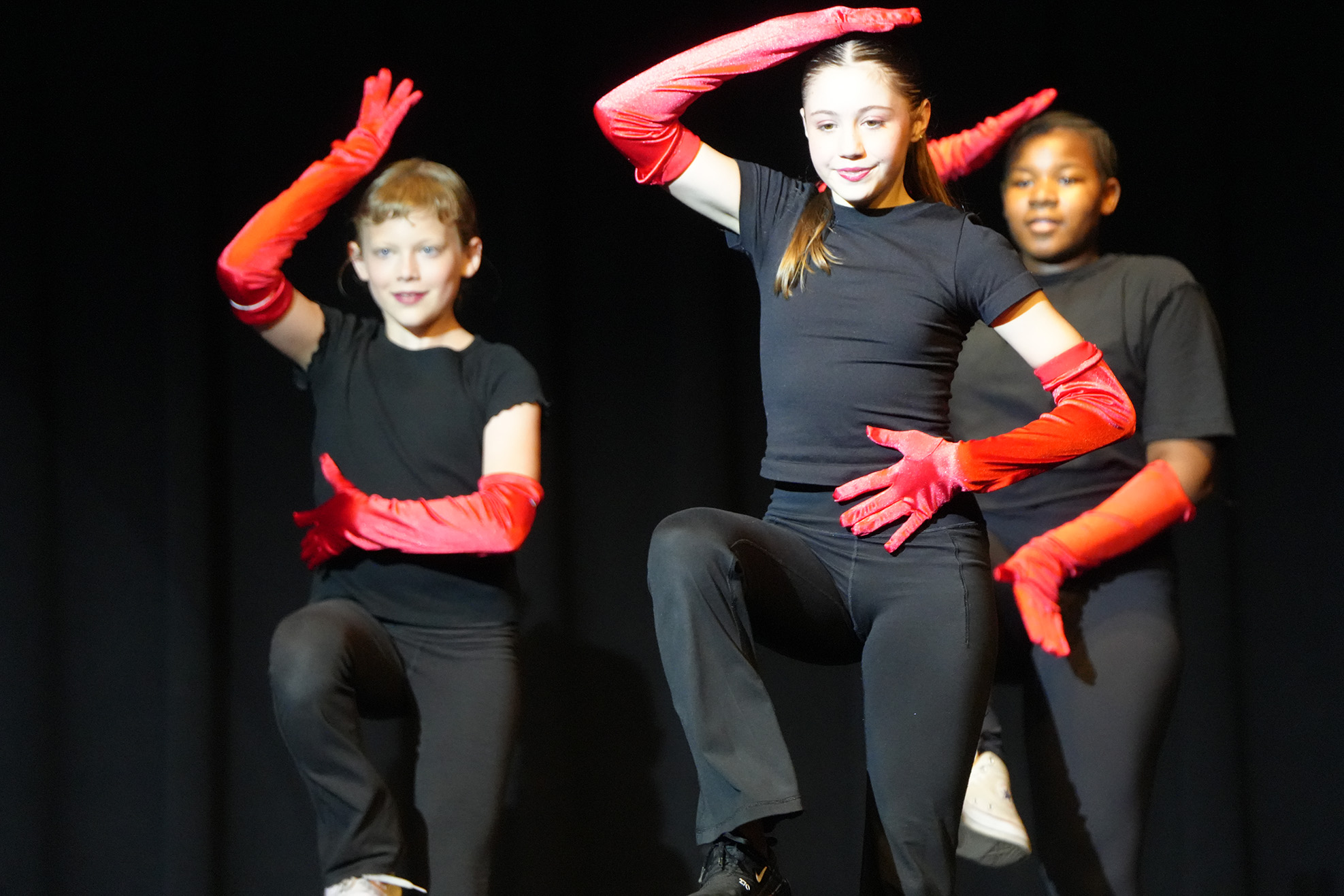 Three children in black t-shirts and red gloves, performing a dance routine on a stage.