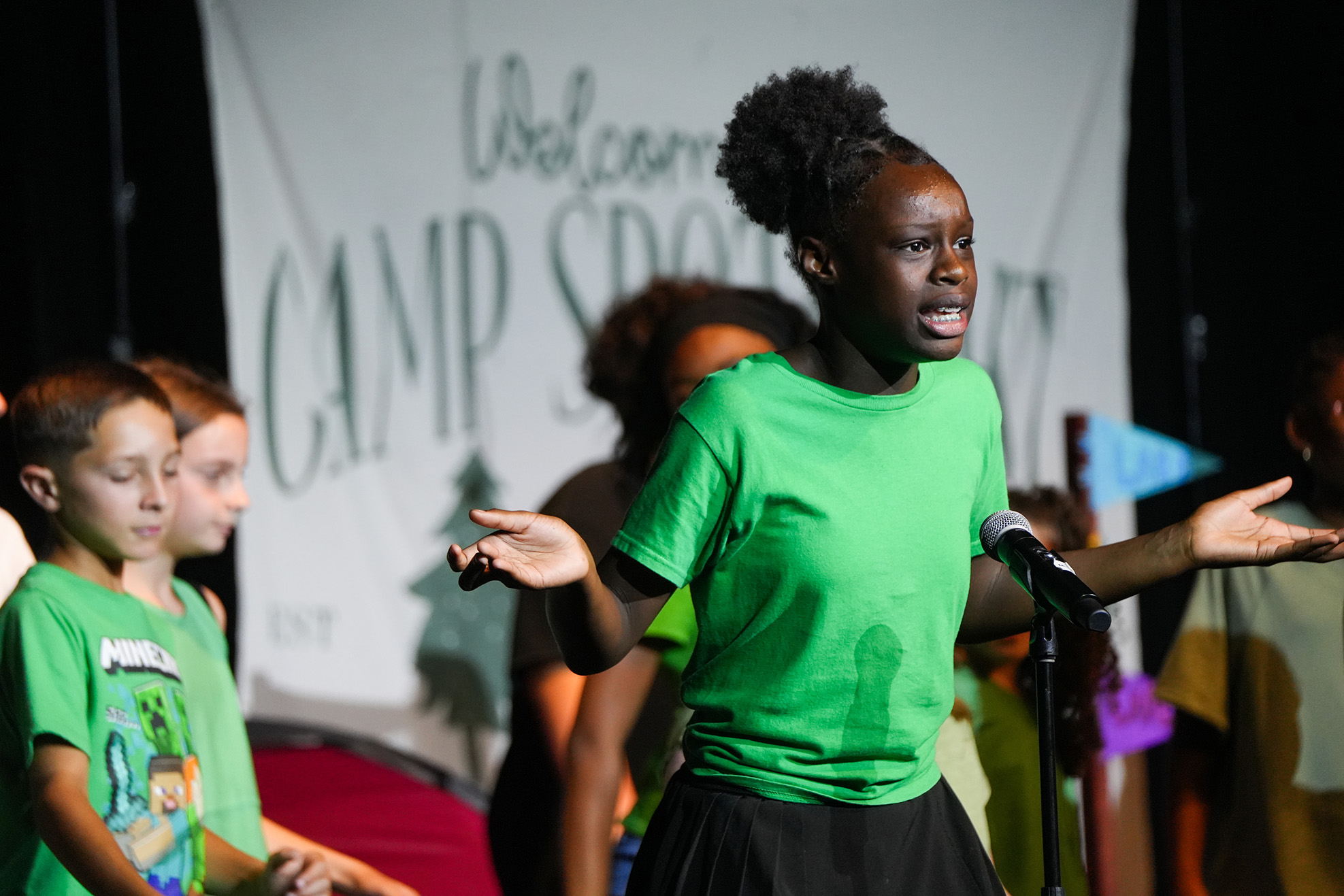 A group of children, one wearing a green t-shirt, standing in front of a banner reading Welcome Camp Sports.