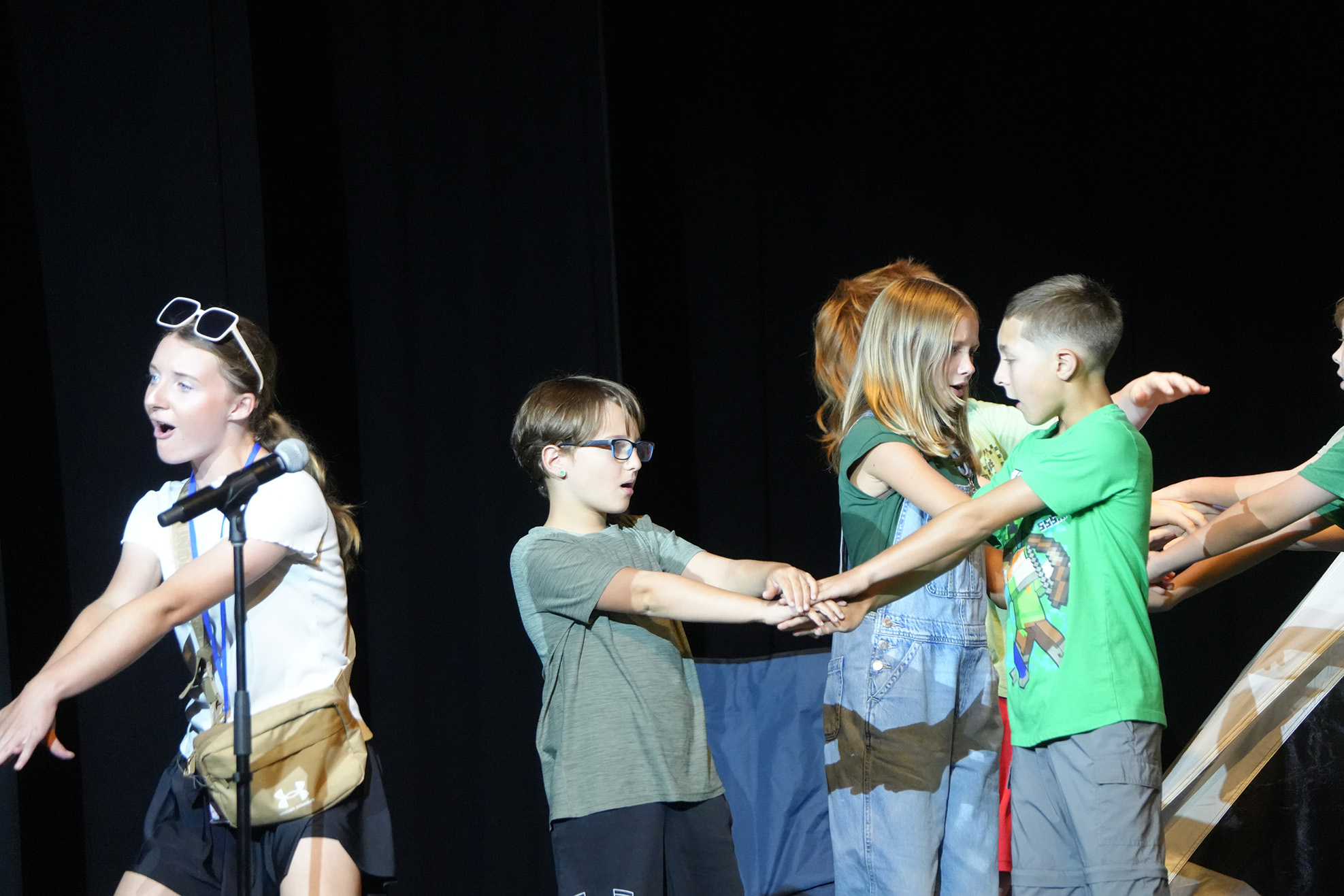 A group of children on stage with one holding a microphone. One child wears glasses, another has sunglasses on their head.