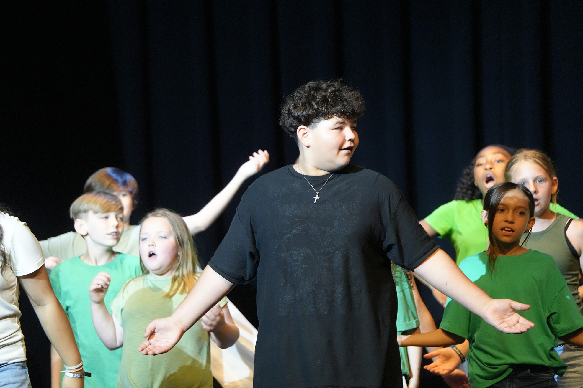 A group of children on stage performing, with one boy in front wearing a black t-shirt.