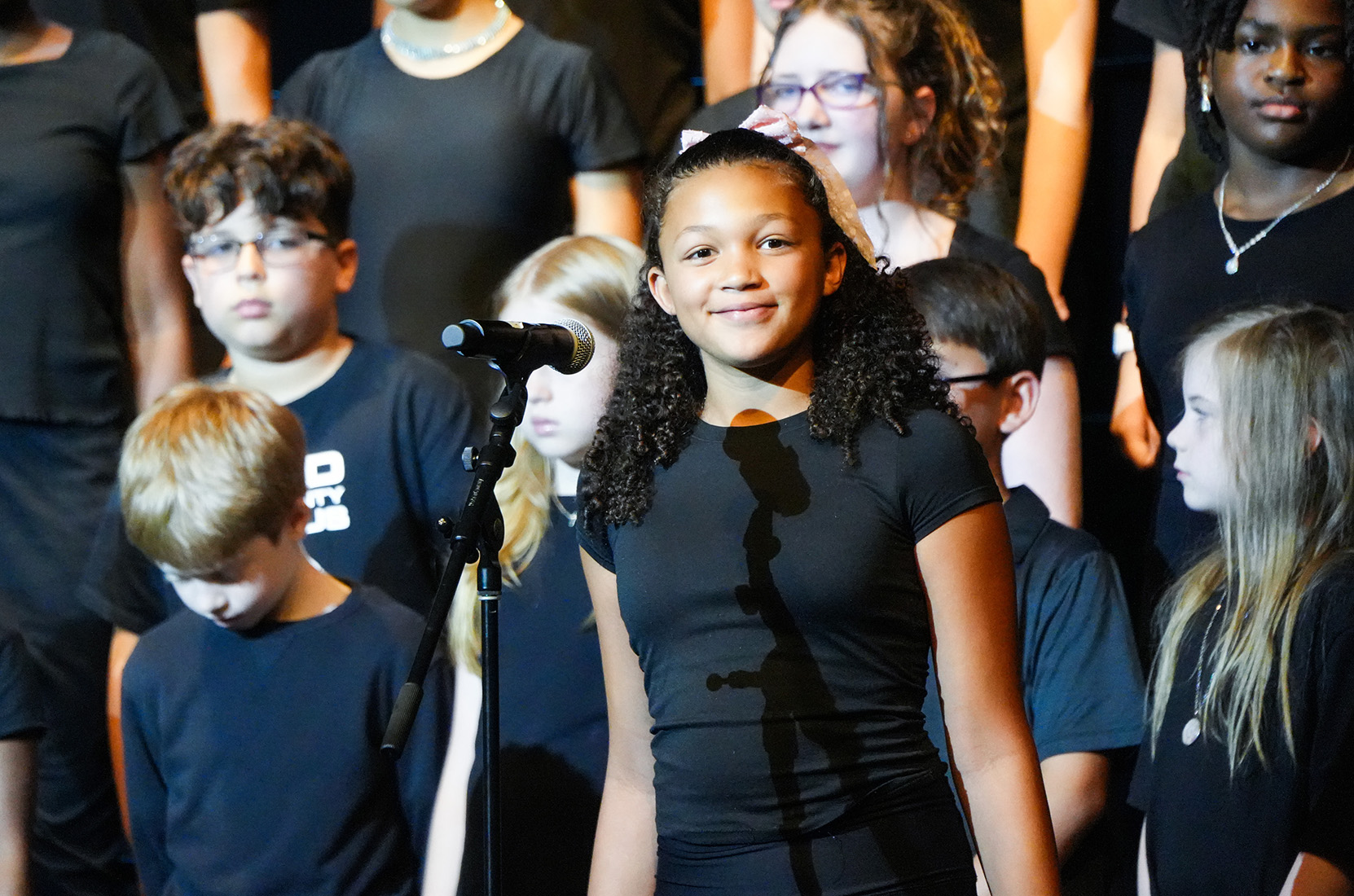 Several children in black t-shirts stand together in a choir; a girl stands in front with a microphone.