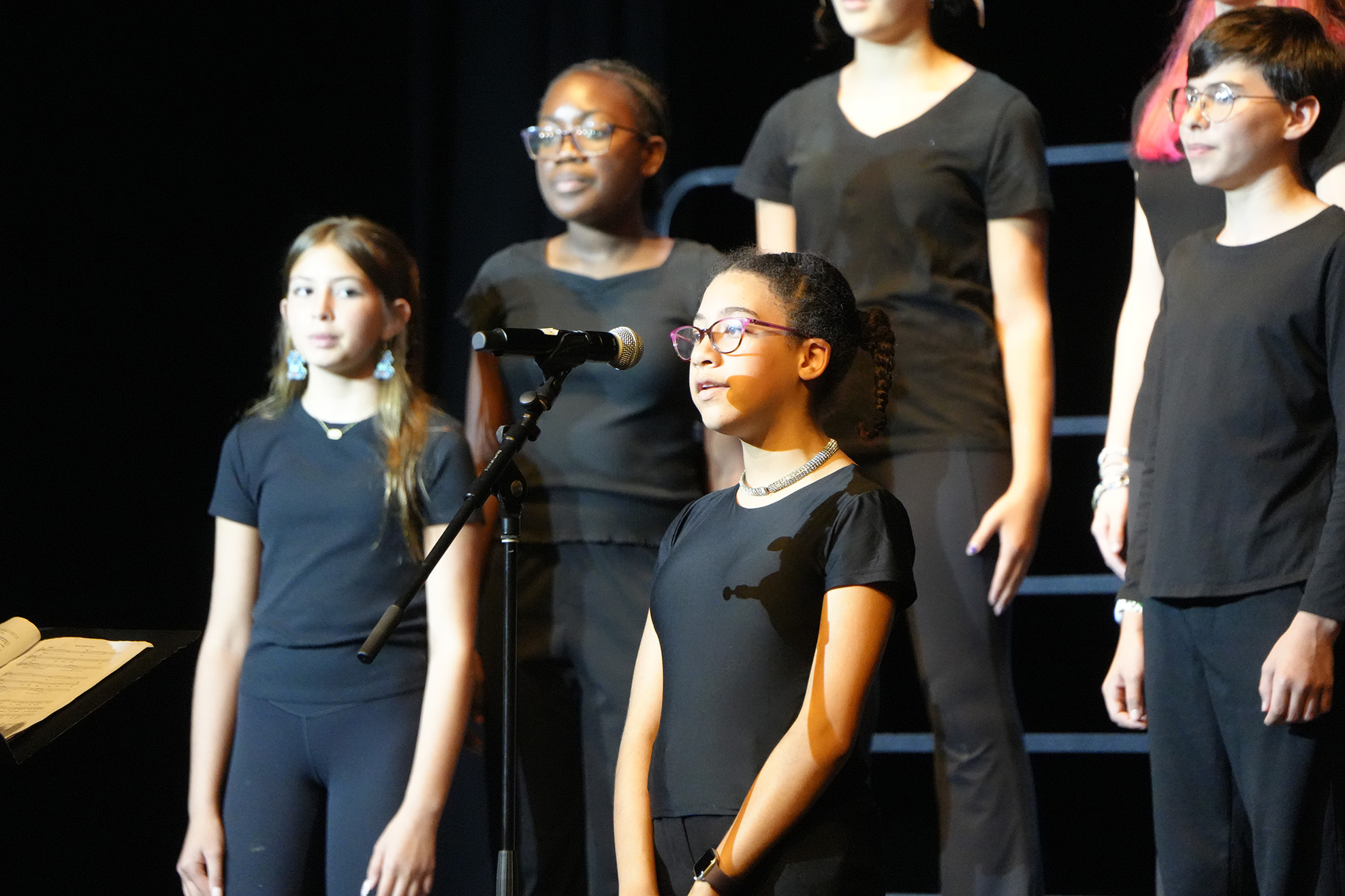 Five children stand on stage in black outfits, with a microphone in front of them. One child stands with a book.