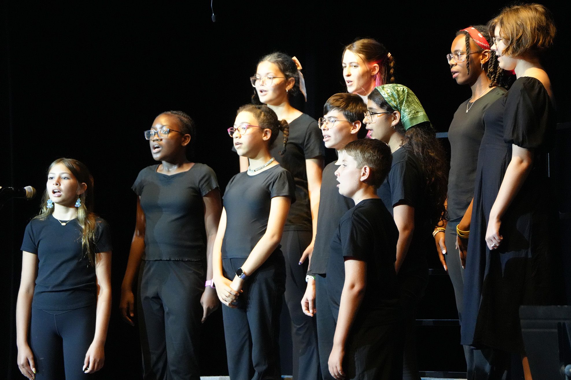A group of young individuals in black attire stand on a stage. They are facing forward and appear to be performing.