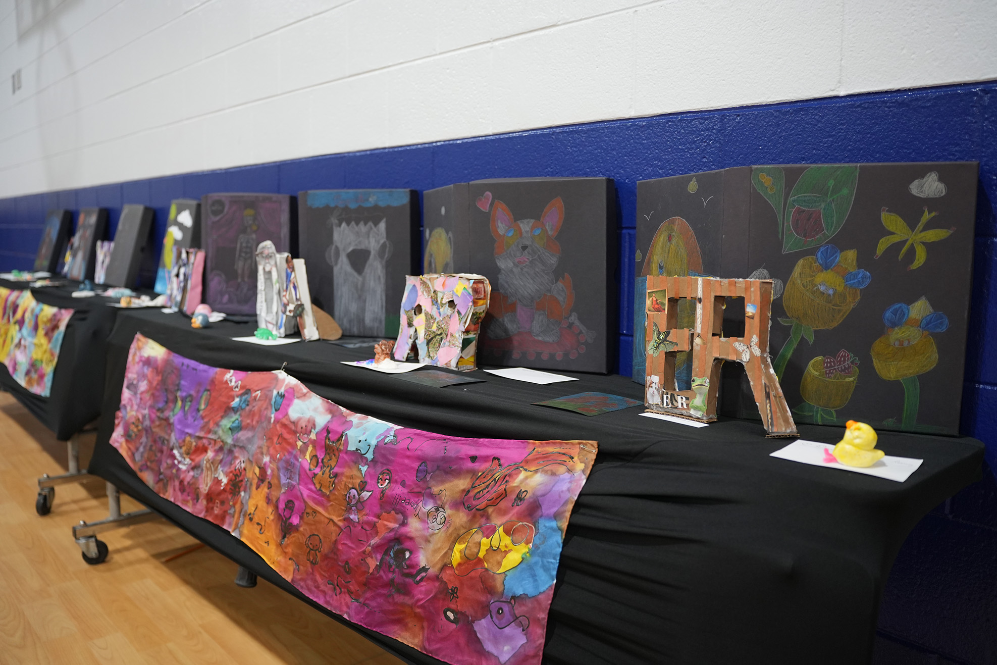 Display of colorful paintings on black tables in a gym. Tables have artworks like cats, letters, flowers, and abstract designs.