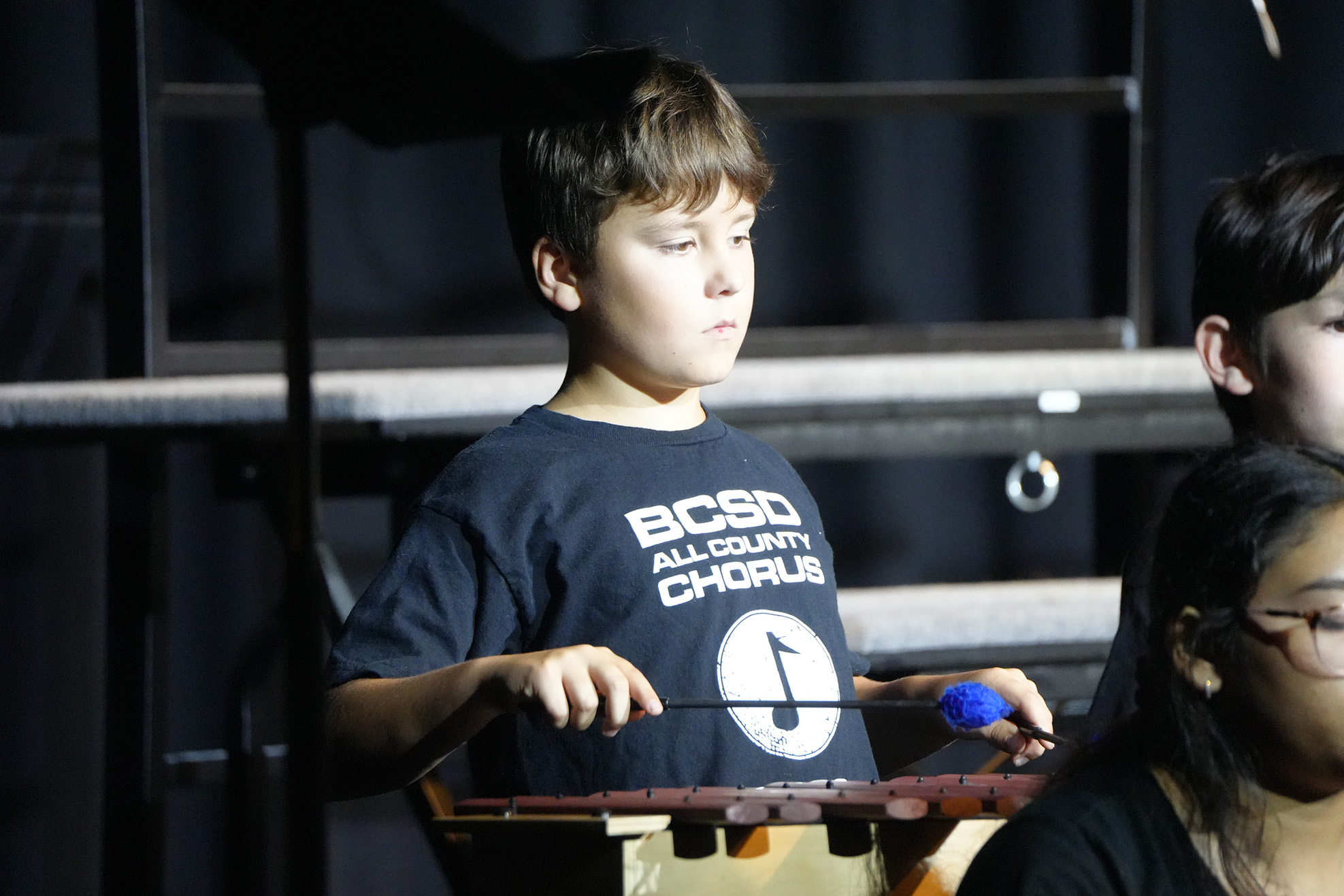 A young boy wearing a "BC80 All County Chorus" shirt plays a xylophone. A girl and another child stand nearby.