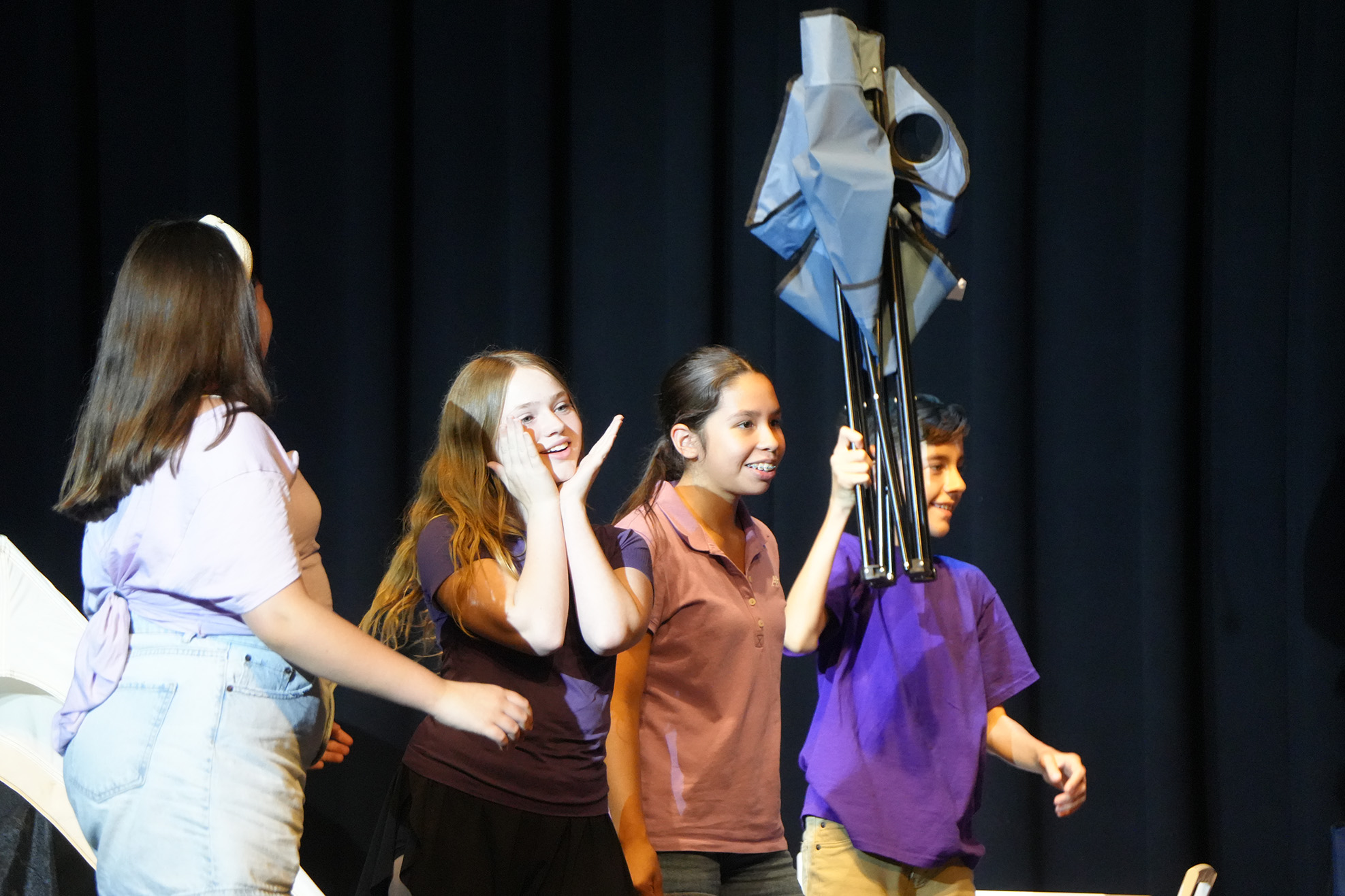 Four children on a stage. Three hold hands and one holds a folding chair. Black curtain in background.