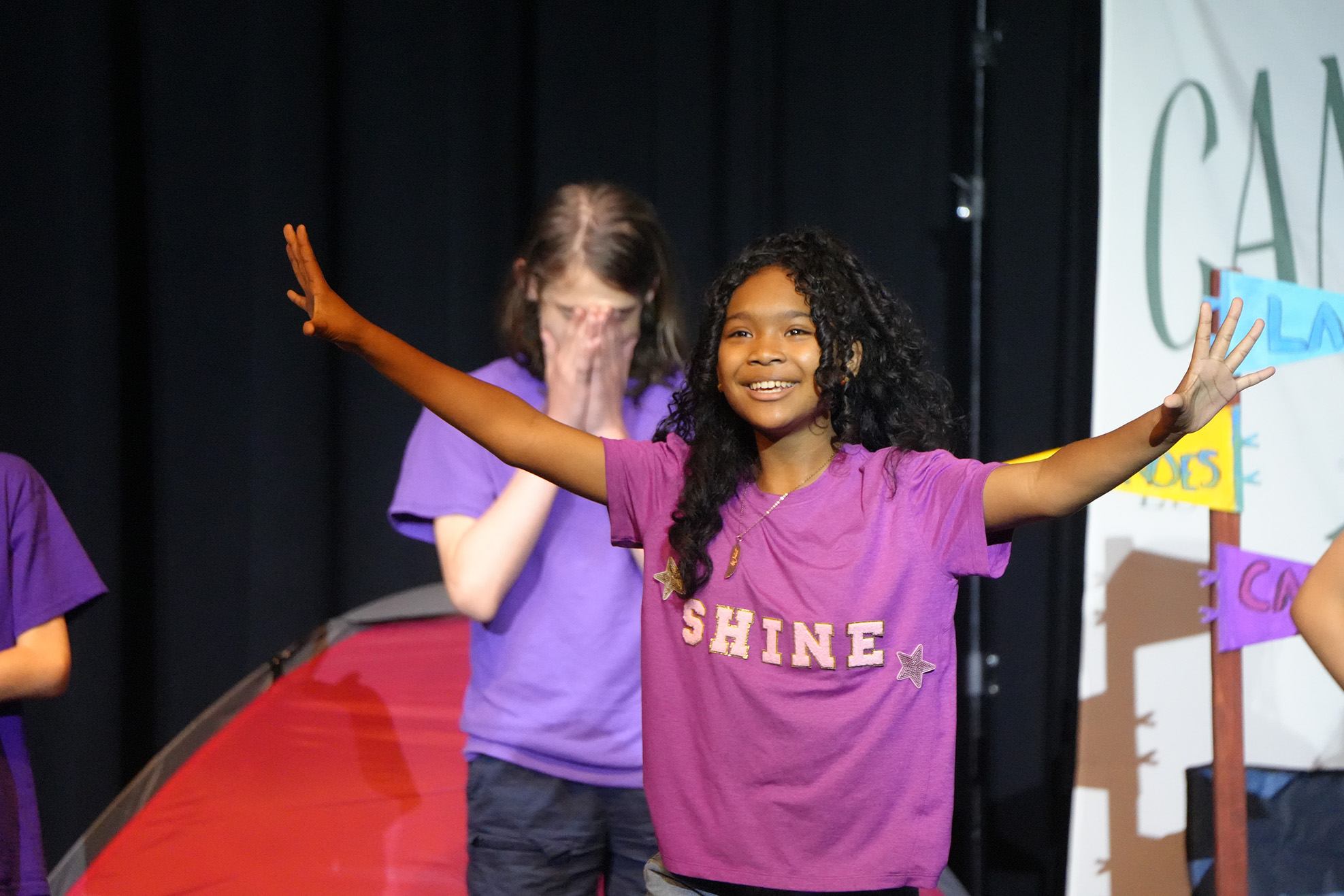 A young person in a purple "Shine" T-shirt with arms outstretched on a stage. Behind, another person in purple stands by a tent.