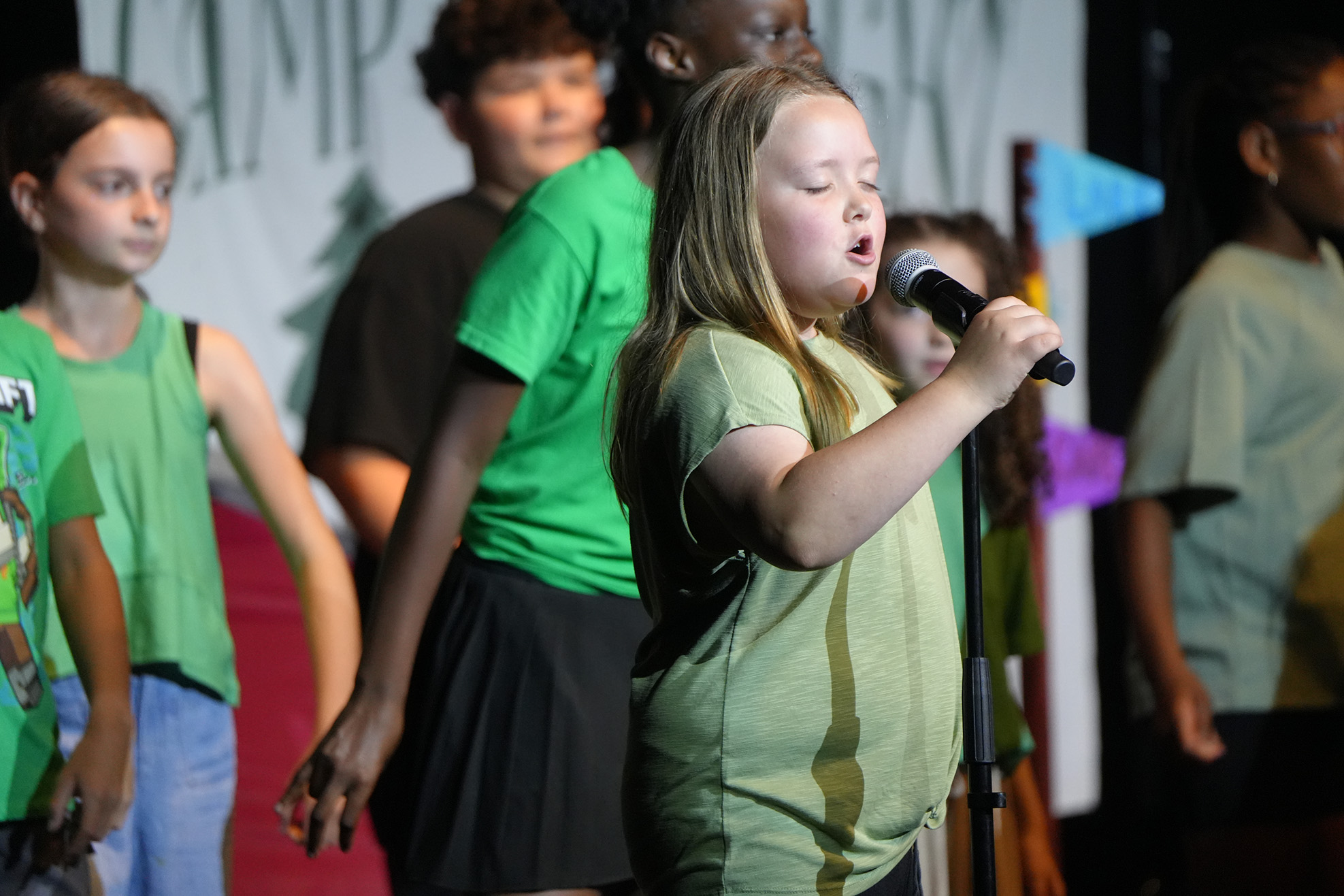 A girl sings into a microphone on stage. Other children stand behind her, all wearing green shirts.