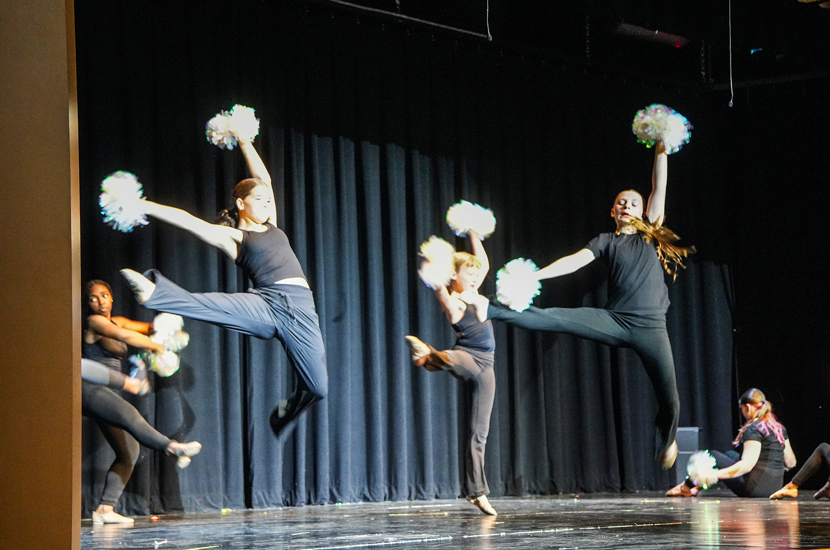 A group of performers in black attire, mid-jump, holding white pom-poms on a stage with a black curtain backdrop.