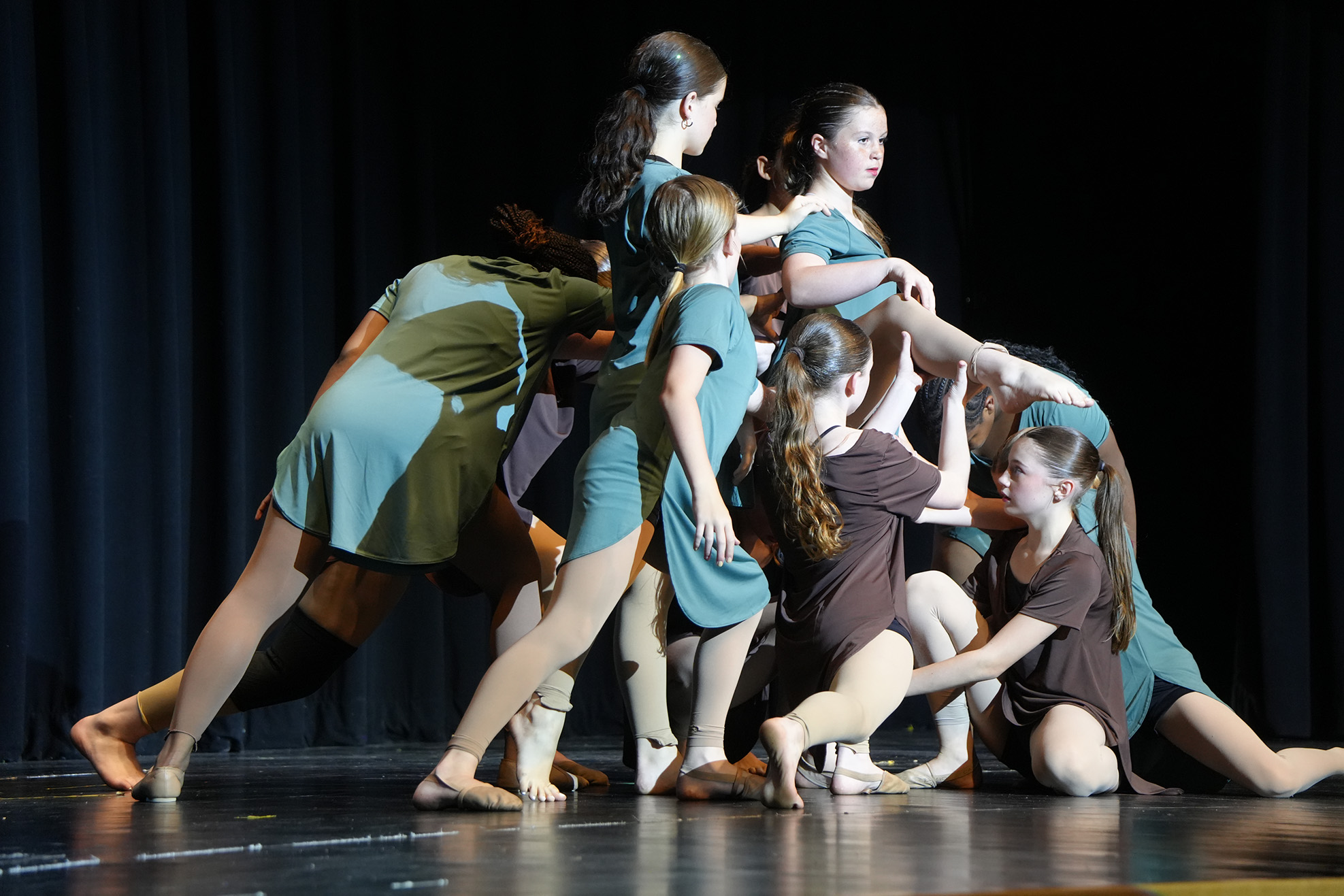 A group of young women in matching dresses perform a coordinated dance on a stage with a black backdrop.