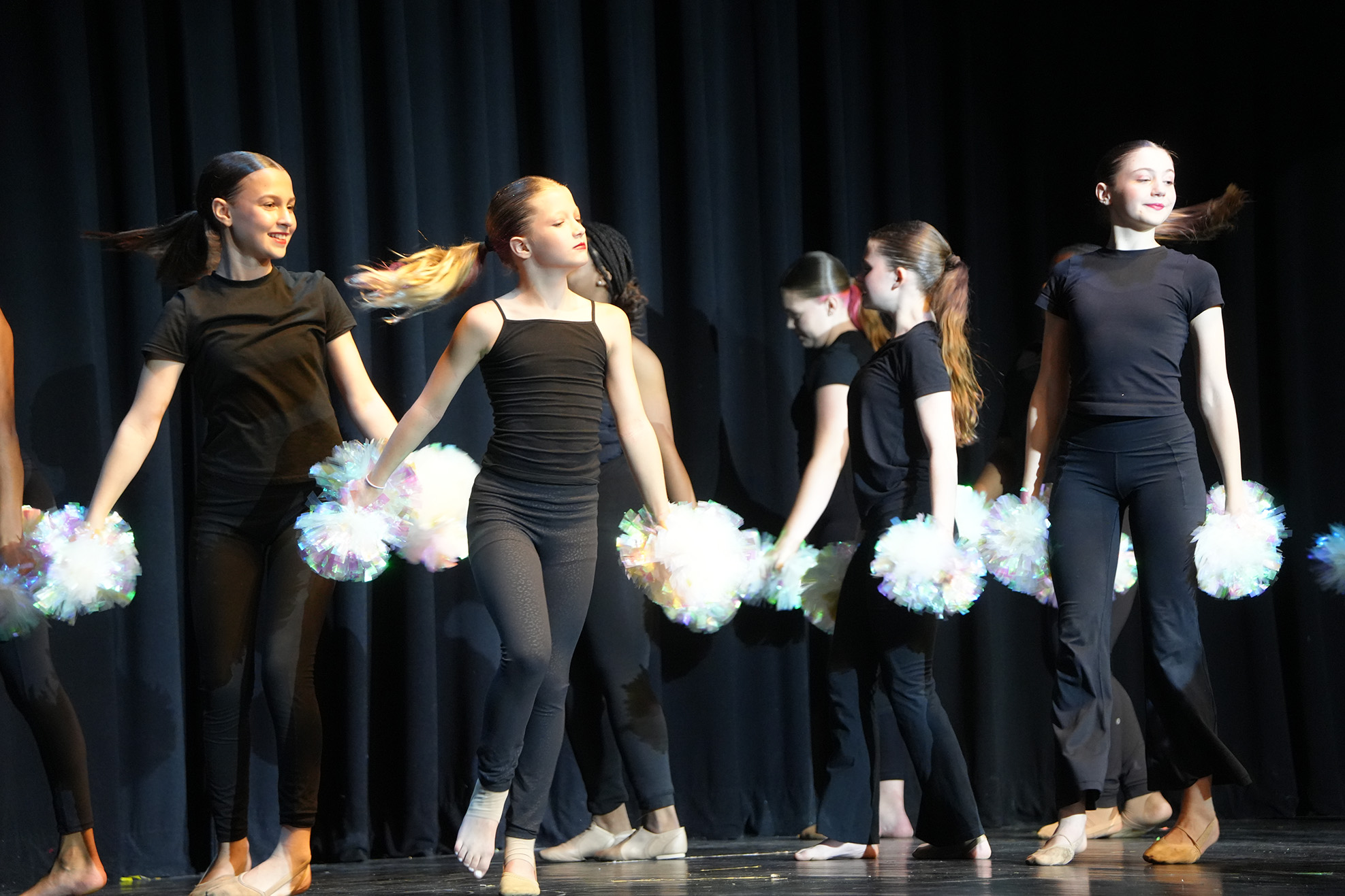 A group of female dancers in black outfits perform on stage, holding white pompoms and executing synchronized dance moves.