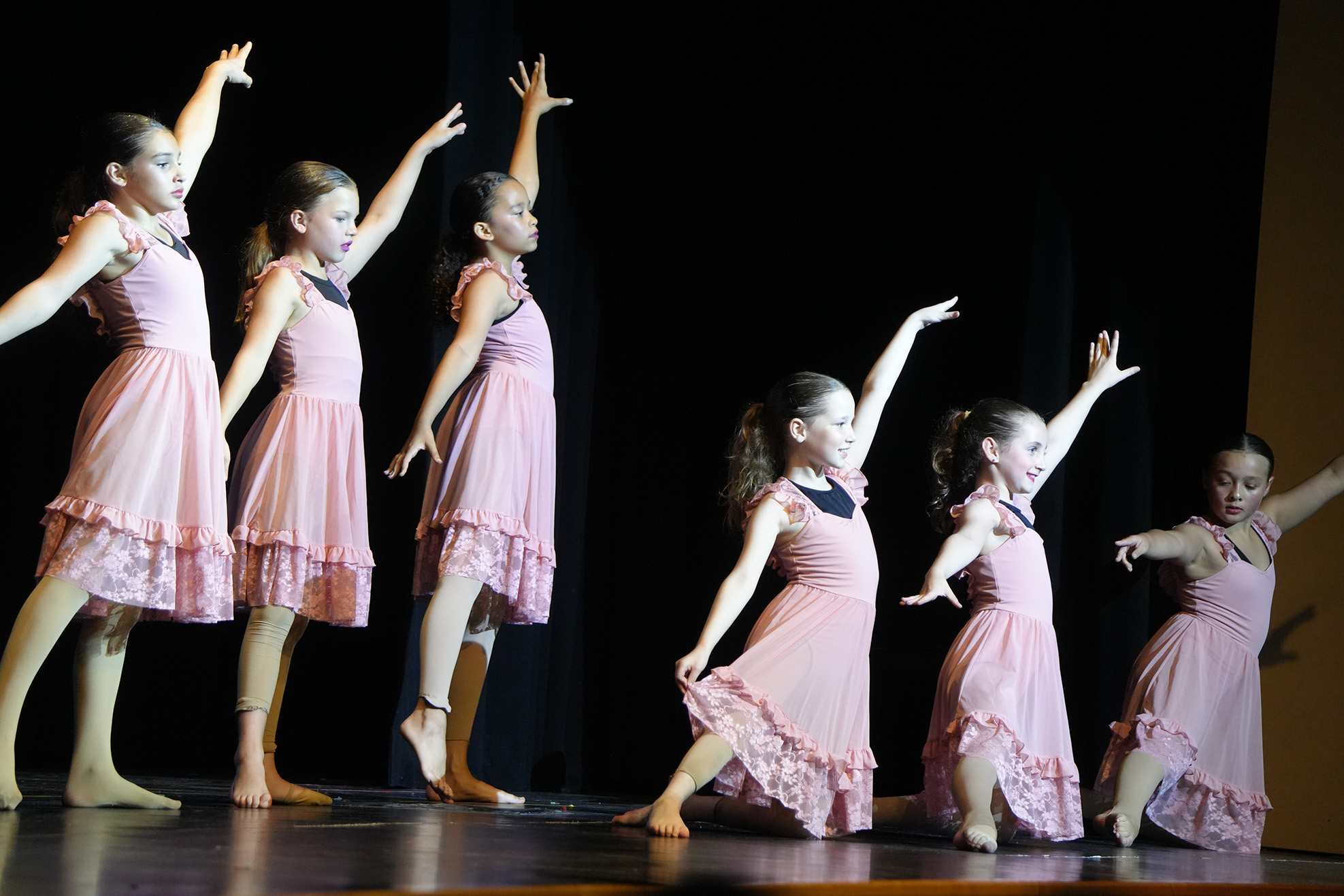 Six girls in pink dresses with ruffles perform on stage, arms and legs extended, against a dark backdrop.