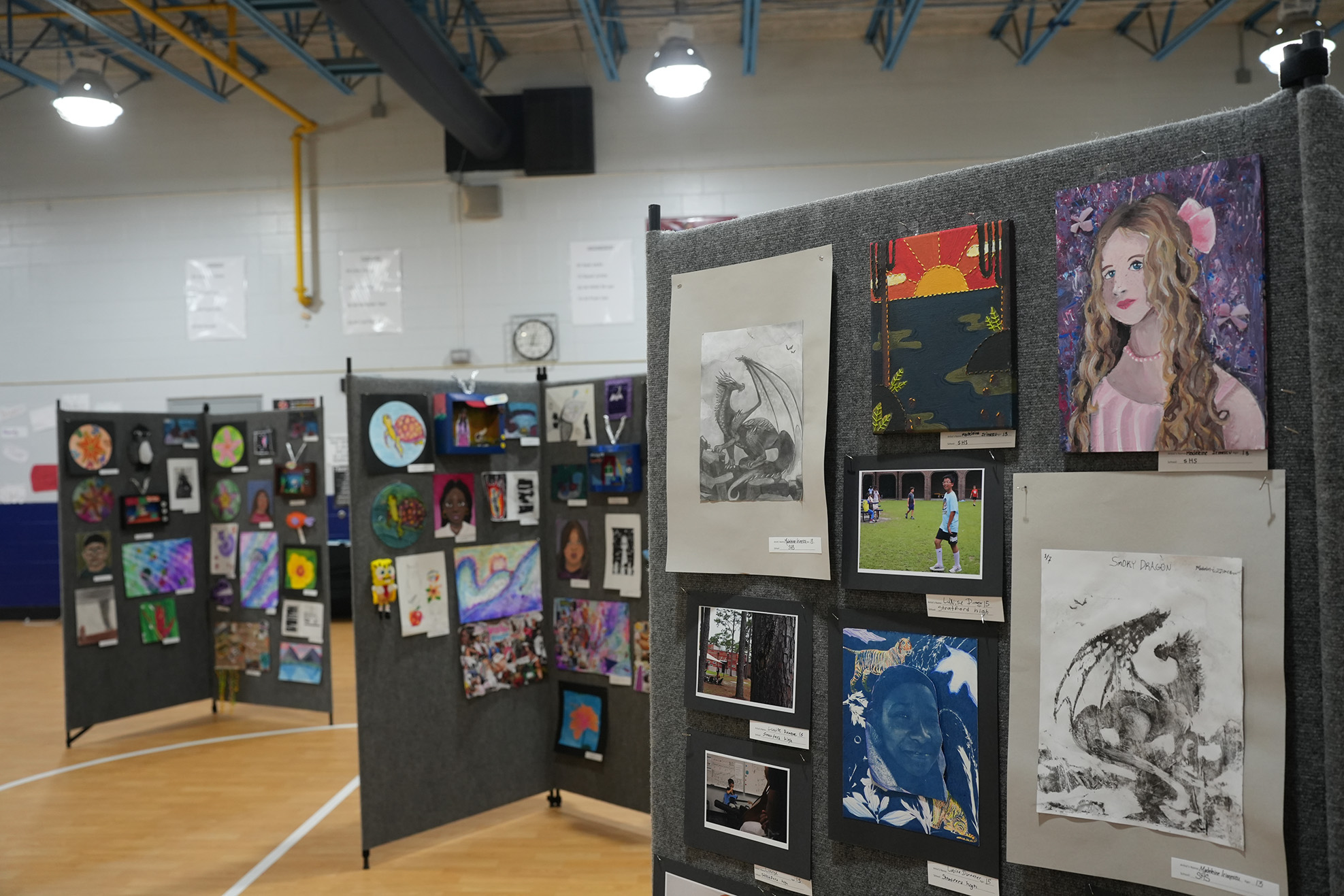 Art exhibition in a gym with diverse artwork on gray display boards. The room has wooden floors and white walls.