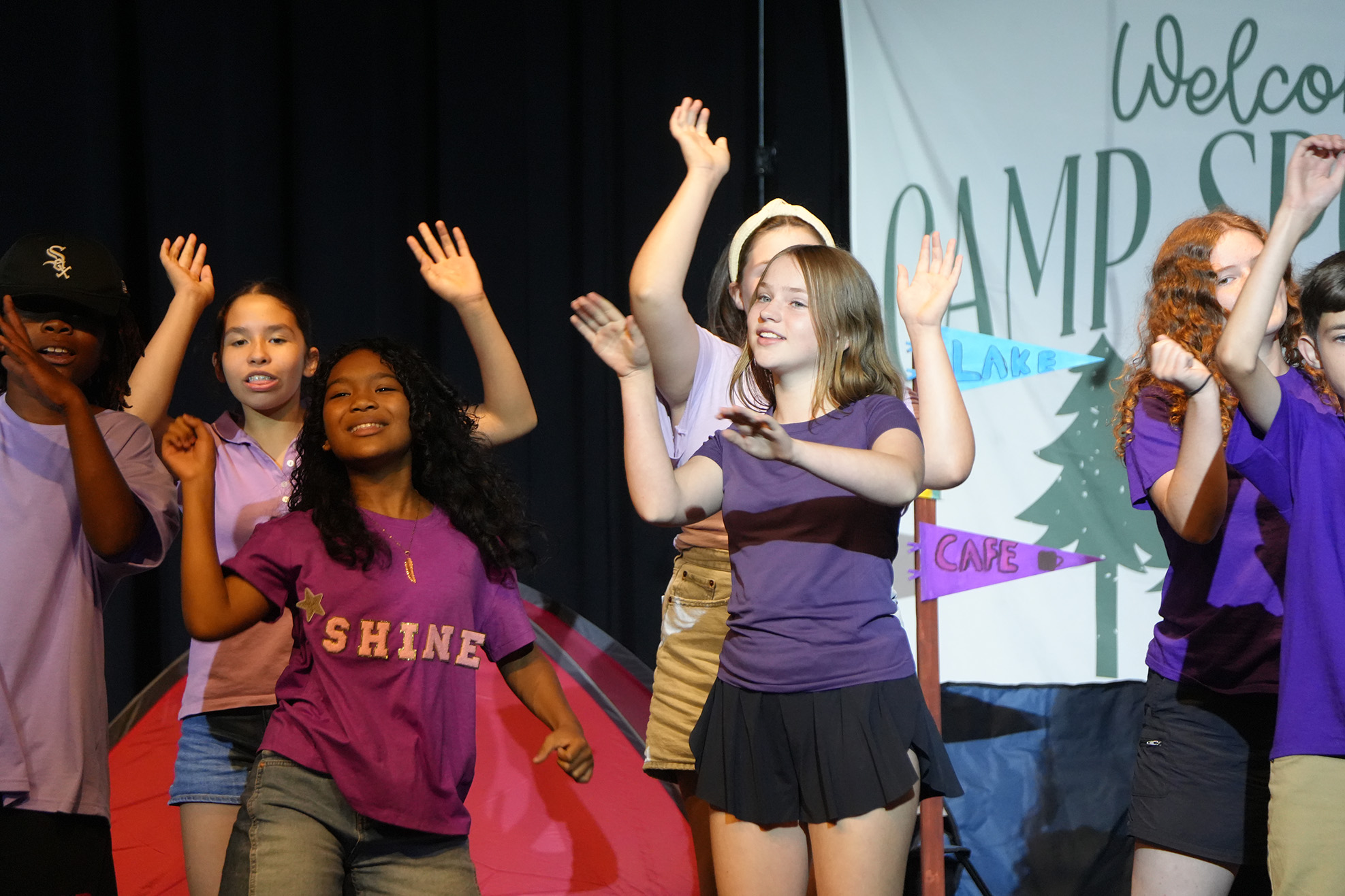 Youth actors perform a skit on a stage. One wears a purple shirt reading "SHINE".
