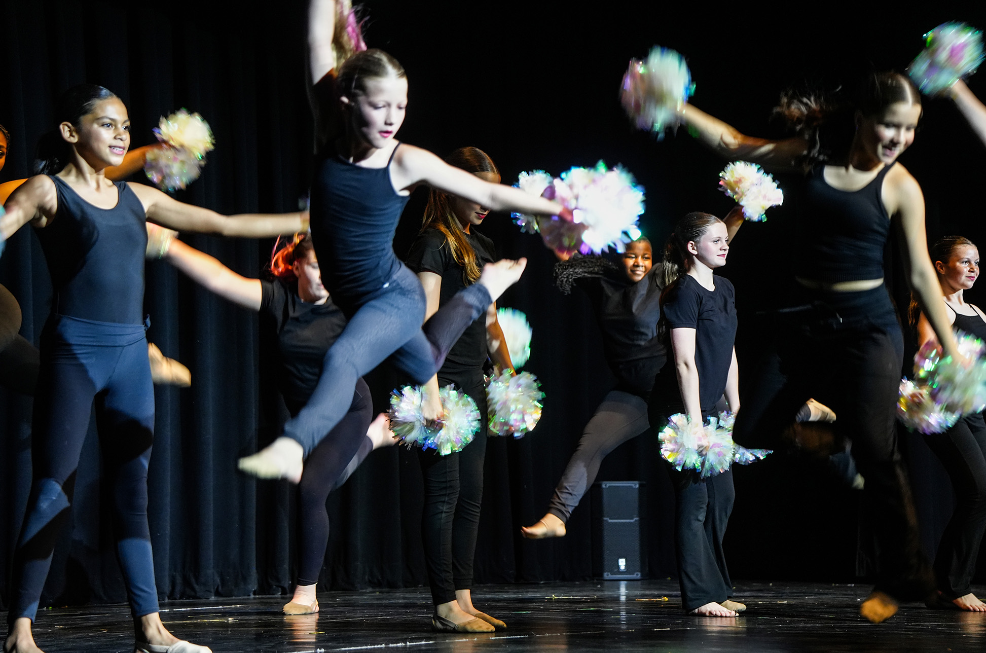 Dancers in black attire perform on stage, holding pom-poms and dancing in a coordinated manner.