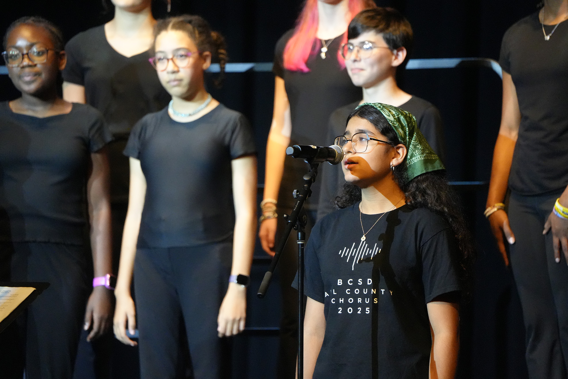 Group of children wearing black clothing, singing into a microphone on a stage with a black curtain backdrop.
