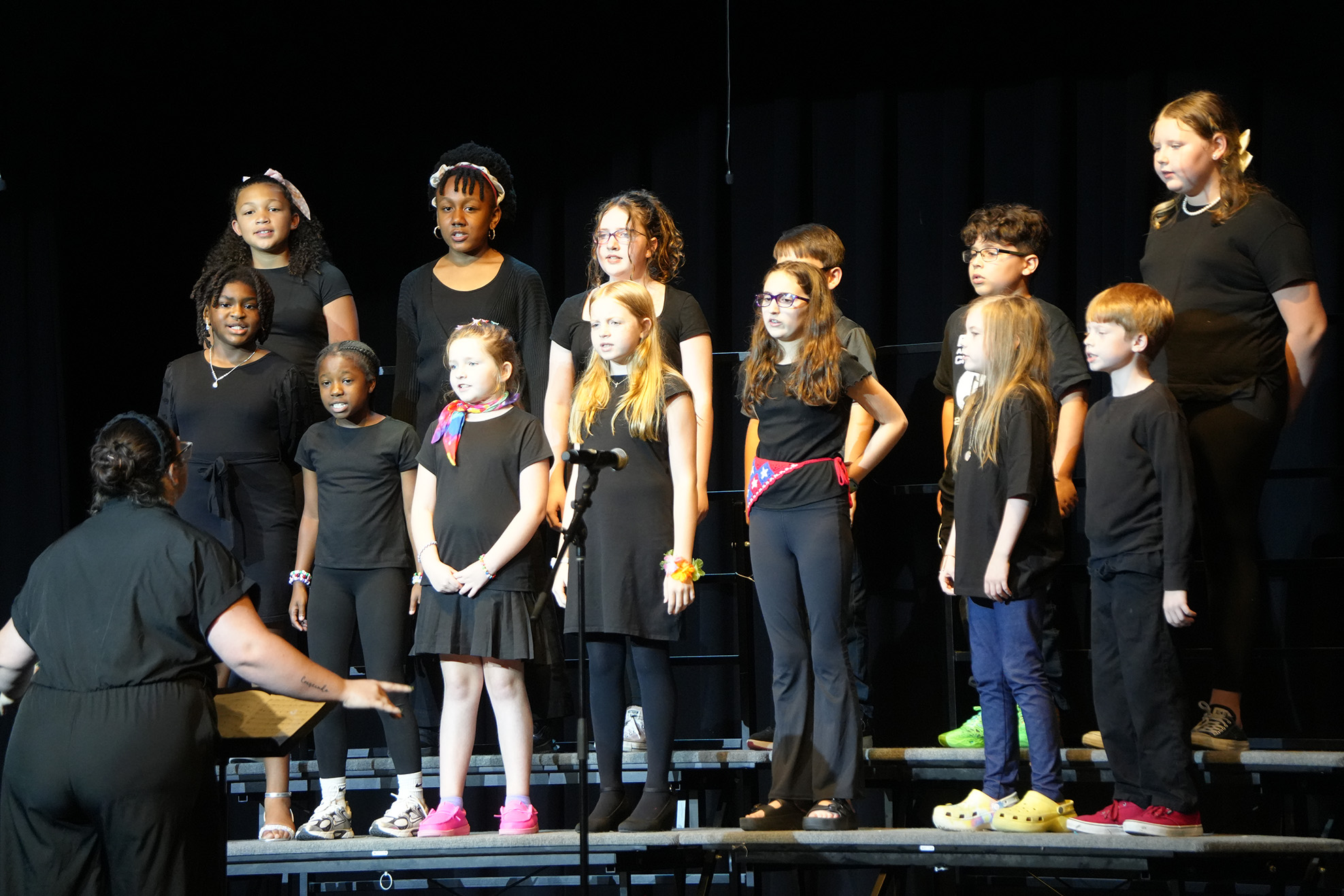 A group of children and adults, all in black, are on stage, singing with a microphone.